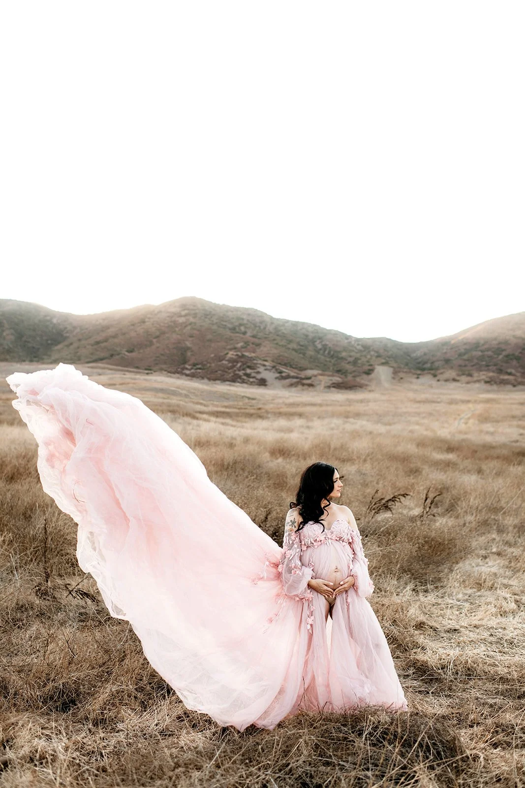 A pregnant woman in a pink flowing dress standing in a dry field with hills in the background.