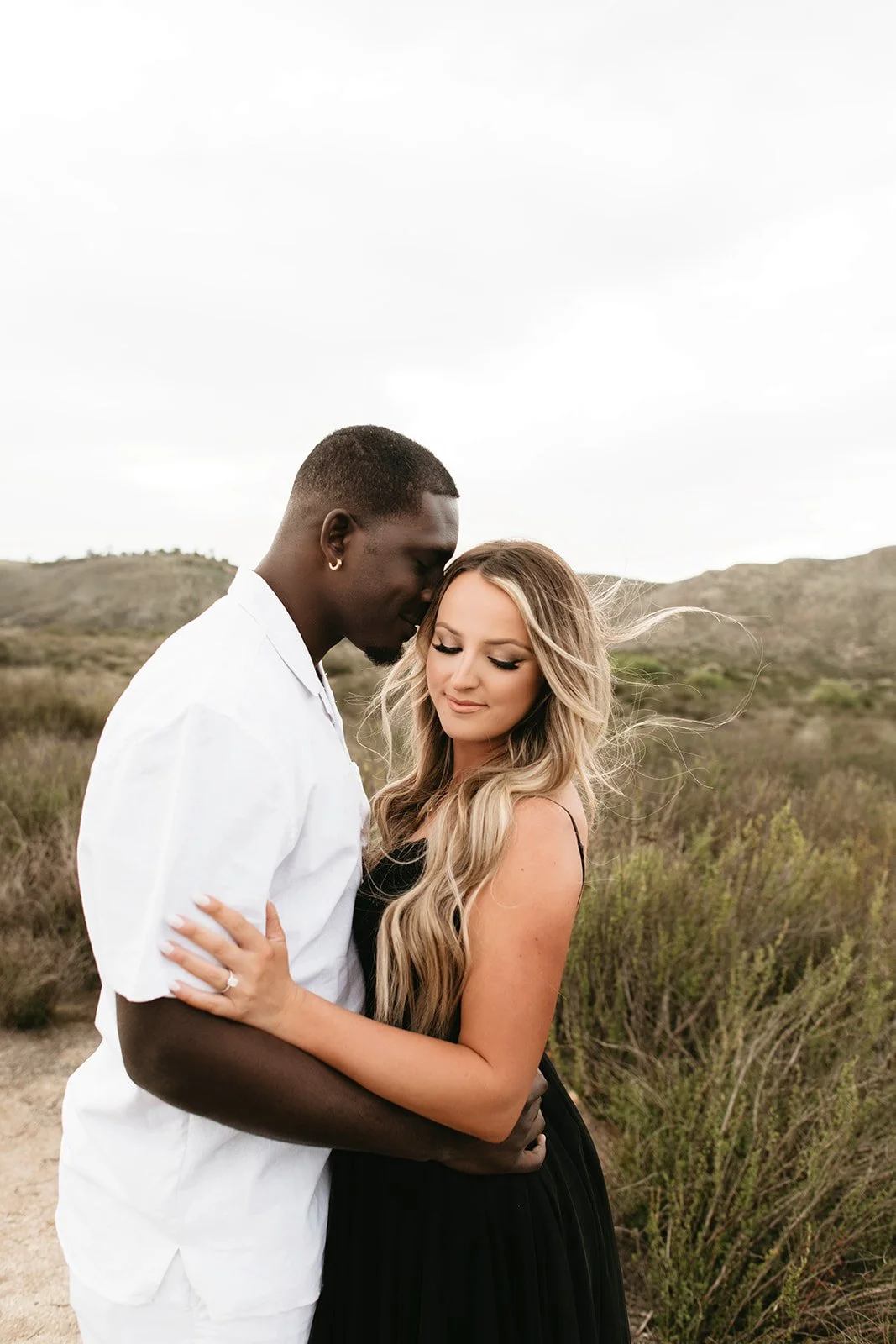 A couple embracing outdoors in a natural setting, with the man gently resting his forehead on the woman's head and both with eyes closed.