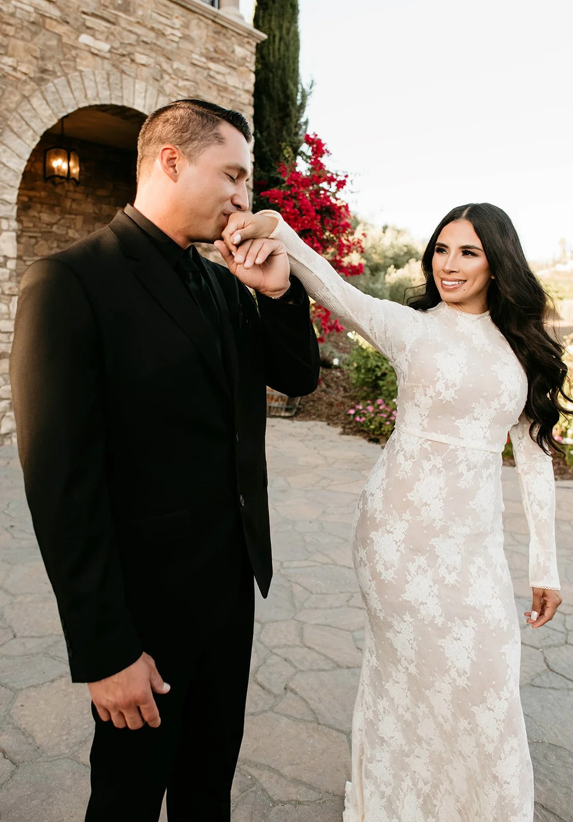 A woman in a white lace wedding dress gently kisses a man in a black suit on the cheek outside a stone building with blooming red and pink flowers in the background.