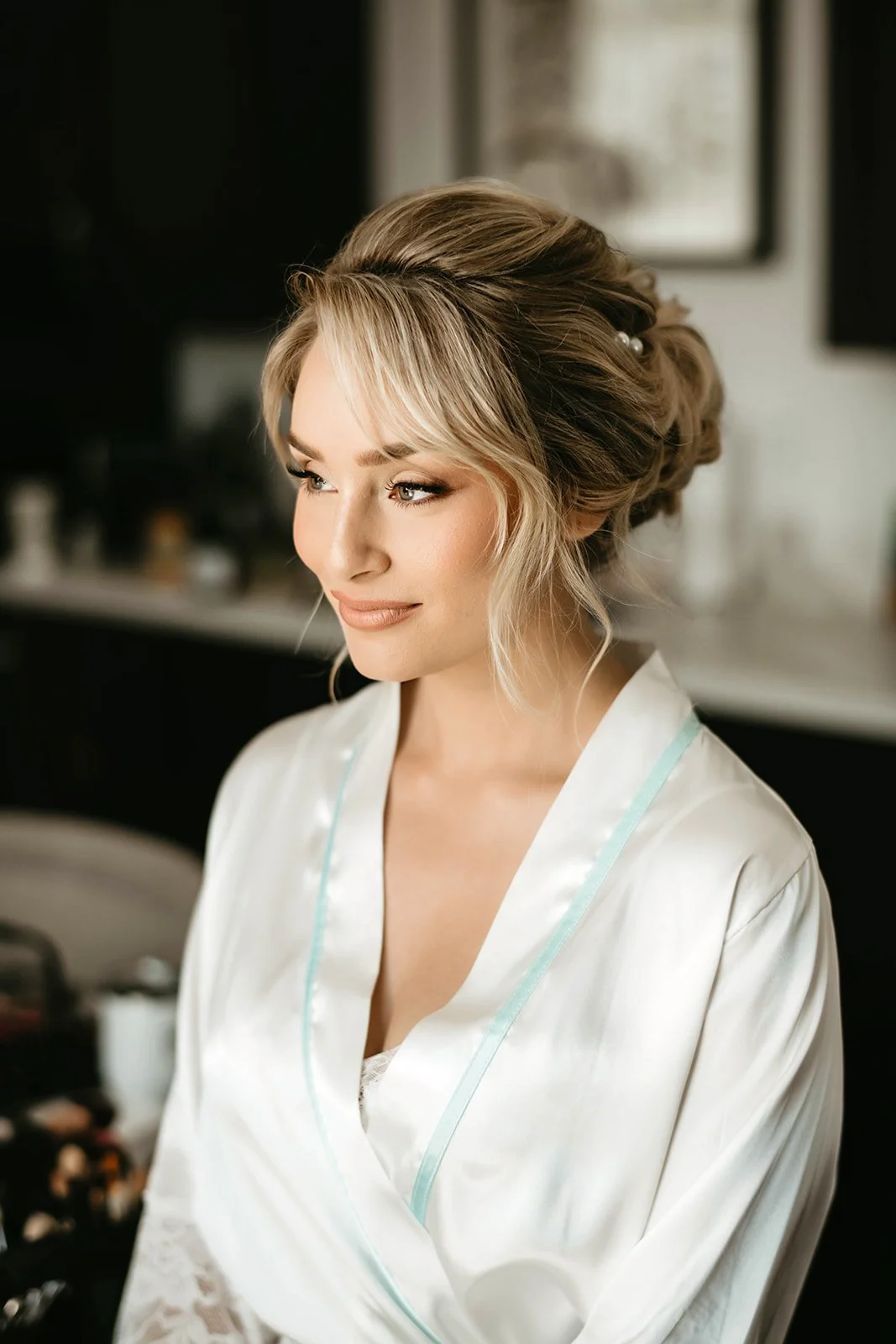 A woman with styled blonde hair, wearing a white satin robe, preparing for her wedding or special event in a softly lit room.