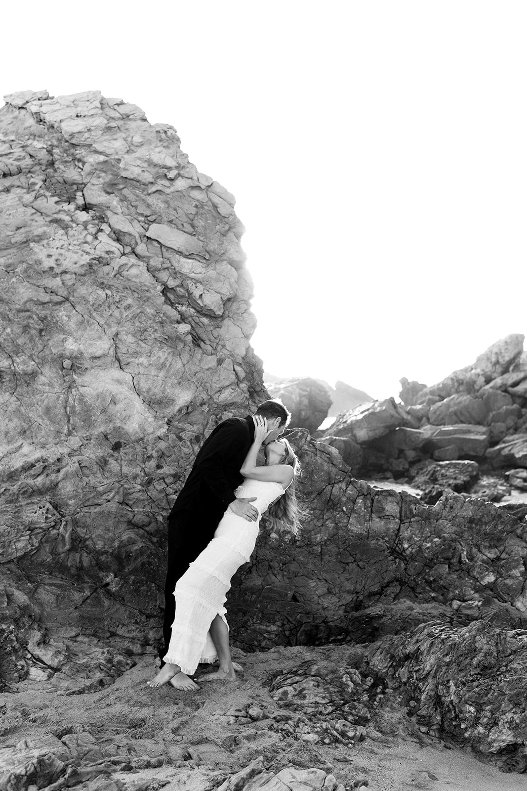 A black-and-white photo of a couple kissing on a rocky beach, the woman in a white dress and the man in dark clothing, with large rocks and cliffs in the background.