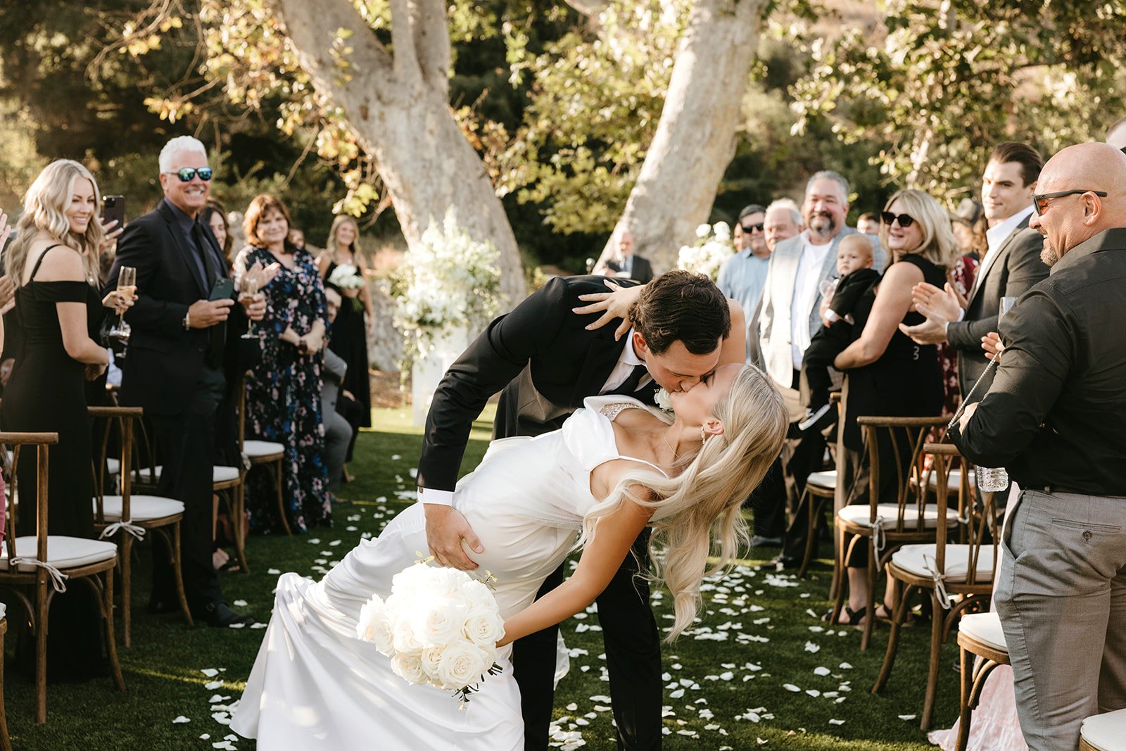A wedding couple shares a kiss during their outdoor ceremony, with guests standing and clapping, surrounded by trees and greenery.