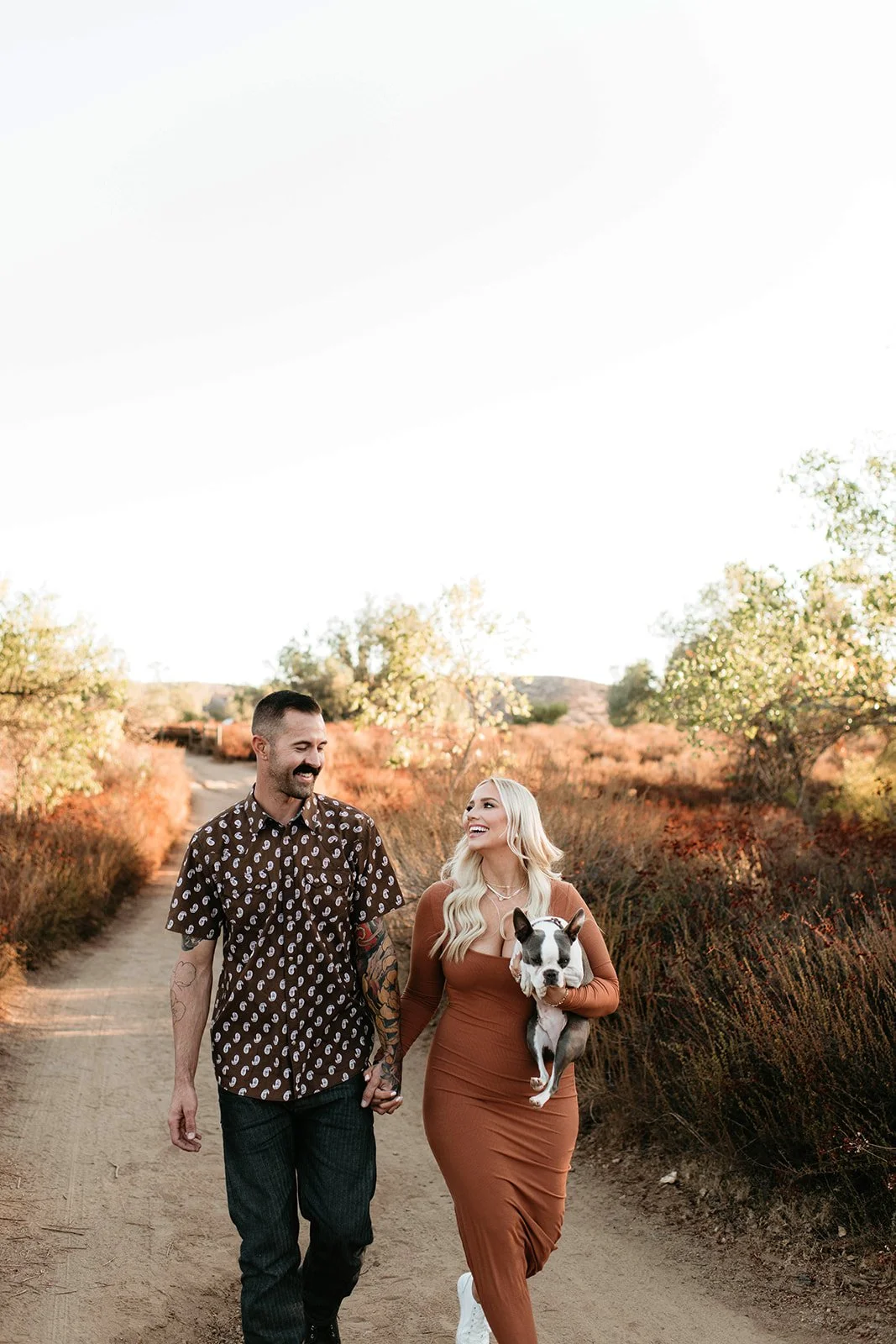 A couple walking down a dirt path holding hands, with a woman holding a small dog, surrounded by autumn foliage.