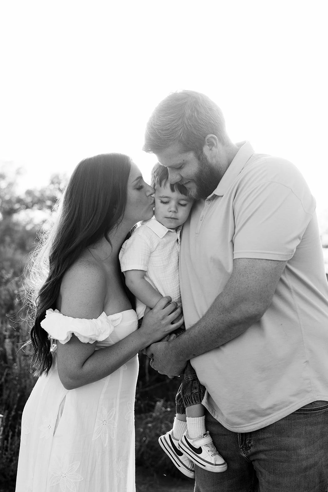 Black and white photo of a family outdoors. The mother and father are holding their young son, with the mother kissing the son's forehead and the father looking down at him. The boy has a neutral expression, and the sunlight creates a bright backgrou