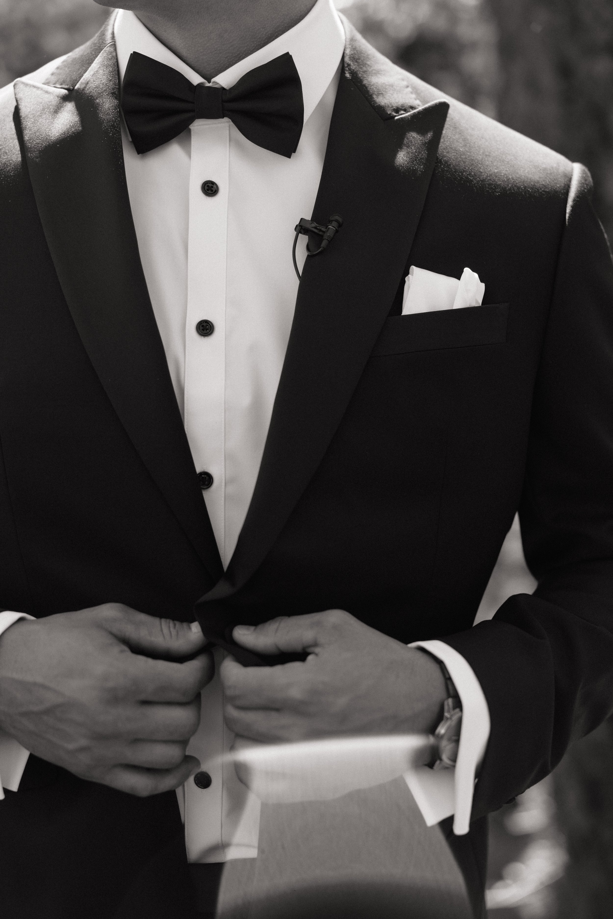 Man dressed in a black tuxedo with a white shirt and black bow tie, holding hands together, with a pocket square visible in the breast pocket.