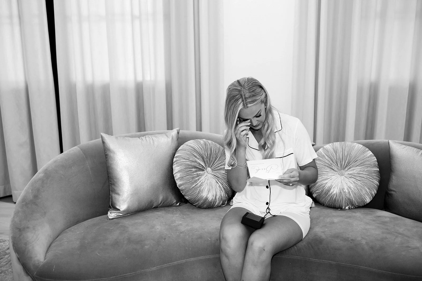 A woman sitting on a curved sofa, holding a piece of paper and a phone, with a distressed expression and tears in her eyes, in a room with curtains.