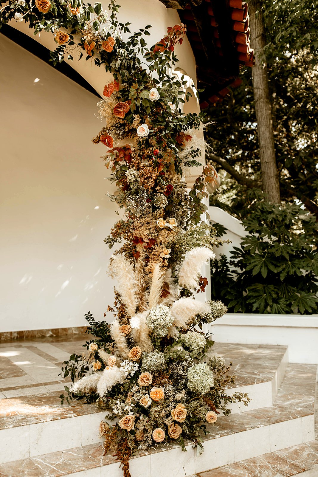 A vertical floral arrangement with cream, white, and peach roses, white baby's breath, and various greenery, placed on marble stairs outside a building with a white wall and trees in the background.