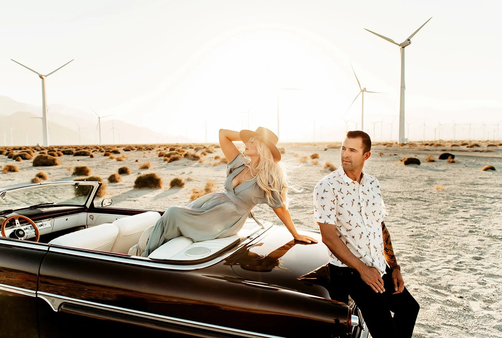 A woman in a flowing dress with a wide-brimmed hat reclining on the hood of a vintage black convertible car in a desert landscape with wind turbines in the background, as a man in a patterned shirt stands beside the car. The scene is sunlit with a cl