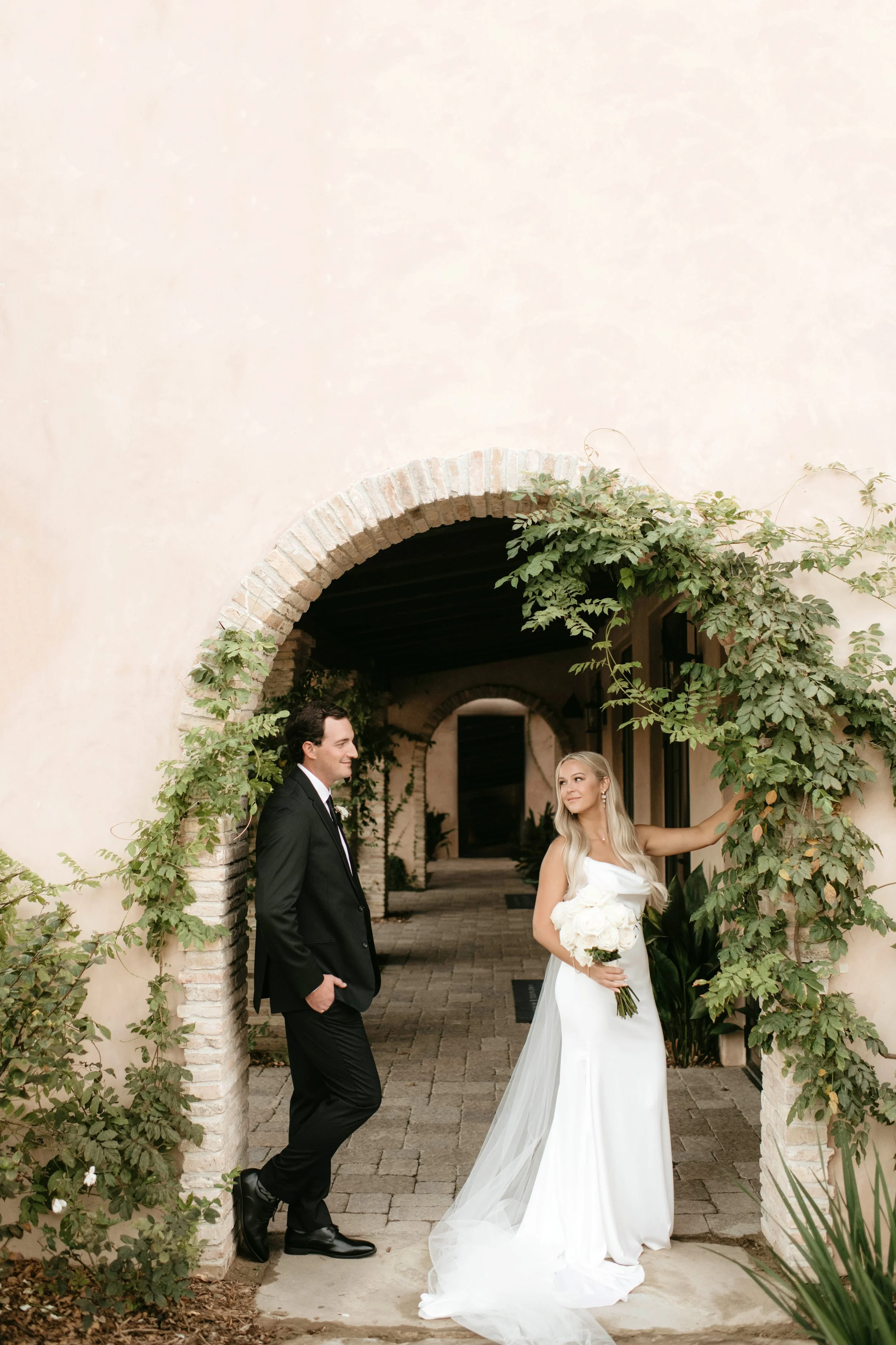 A bride in a white wedding dress holding a bouquet of white flowers, and a groom in a black suit smiling at her, standing under an arched brick entrance with green foliage.