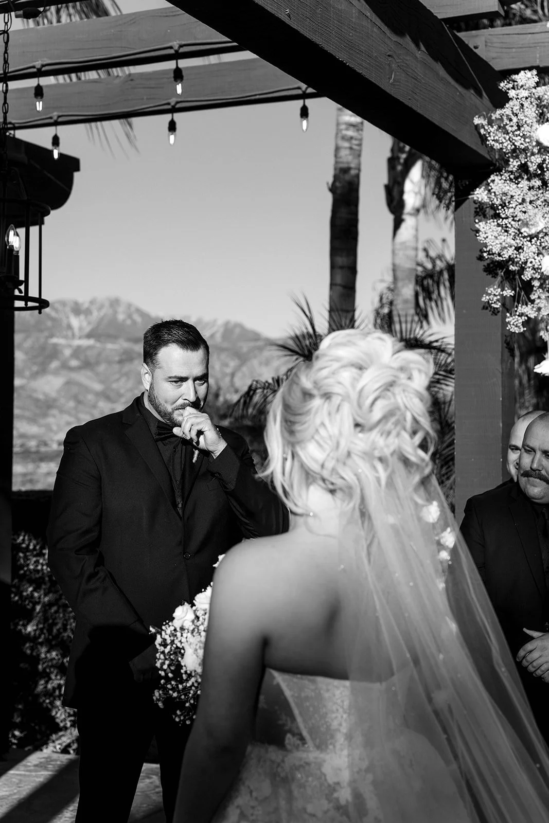 A black and white photo of a wedding ceremony outdoors with mountains in the background. The groom, in a dark suit, is emotional and appears to be wiping away tears, while the bride, with her back to the camera, has styled blonde hair with flowers an