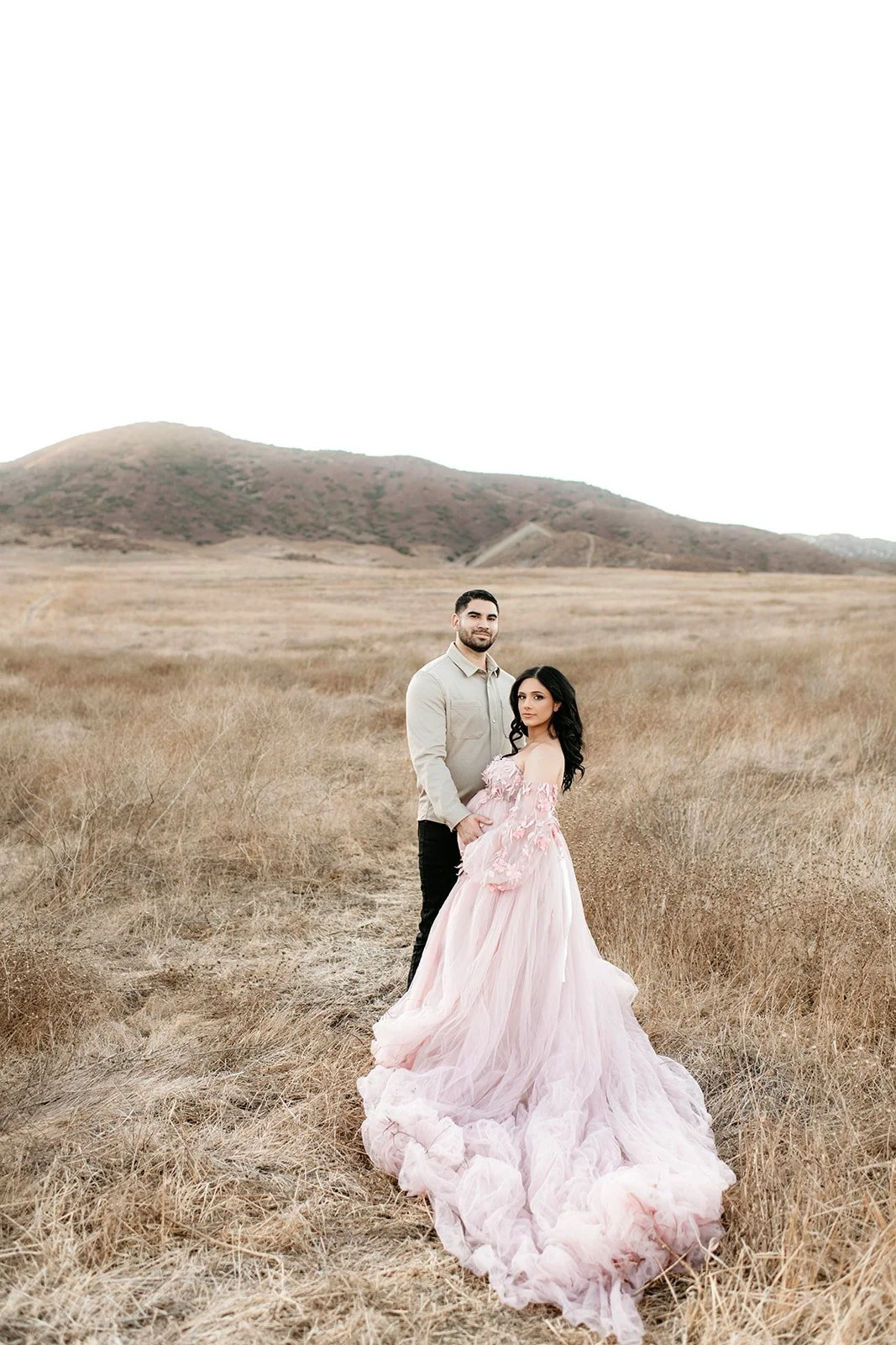 A couple standing in a dry grassy field with a mountain in the background, the woman is wearing a pink, off-shoulder, flowing gown, and the man is dressed in a beige shirt and black pants.