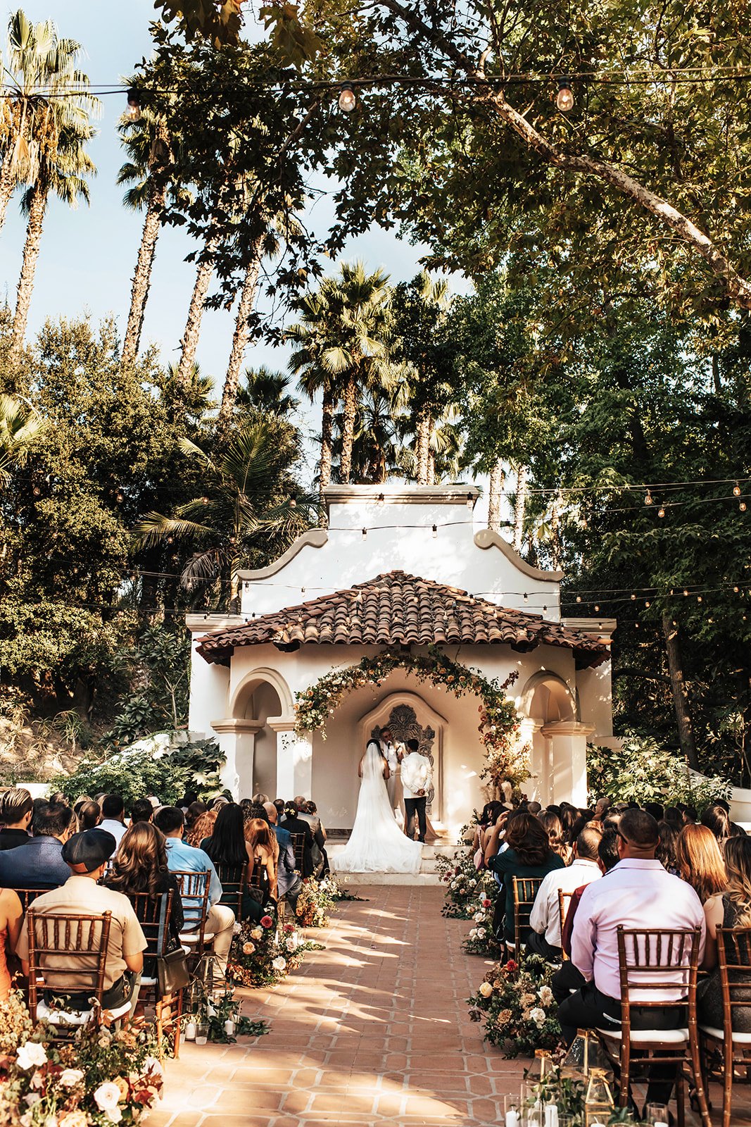 Outdoor wedding ceremony with guests seated facing a small white chapel decorated with floral arrangements, under tall palm trees and string lights.