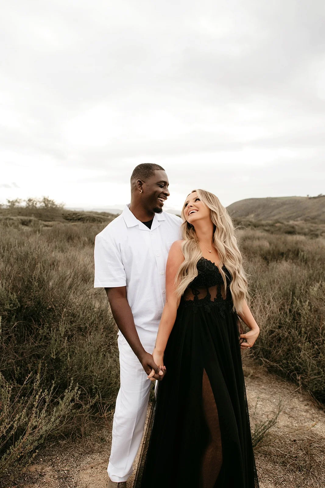 A happy couple holding hands and smiling at each other in a grassy outdoor landscape under a cloudy sky.
