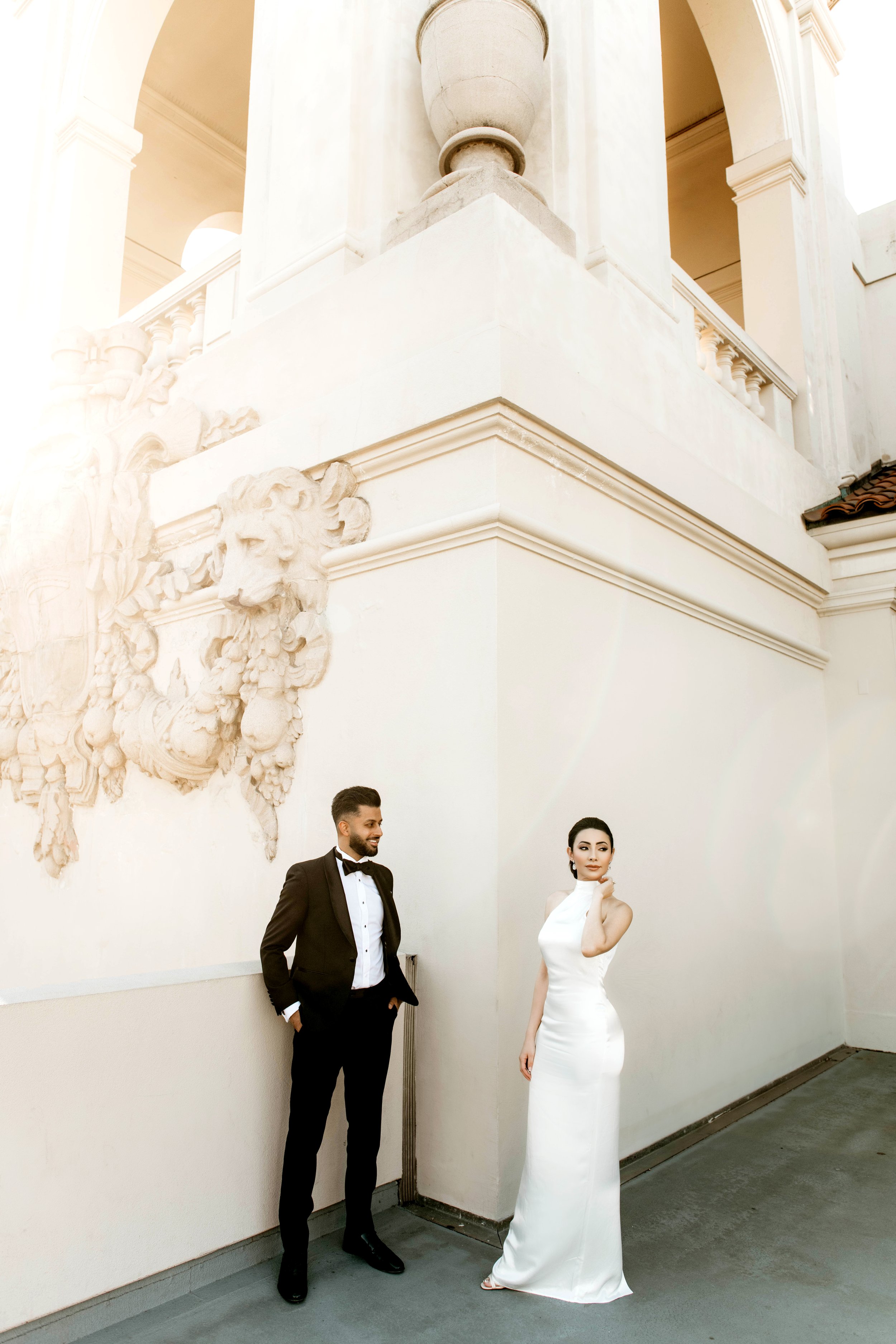 A man in a black tuxedo and a woman in a white sleeveless gown stand against a white wall decorated with a carved lion's head sculpture and ornate architectural details. The man has short dark hair and a beard, smiling with hands in pockets. The woma