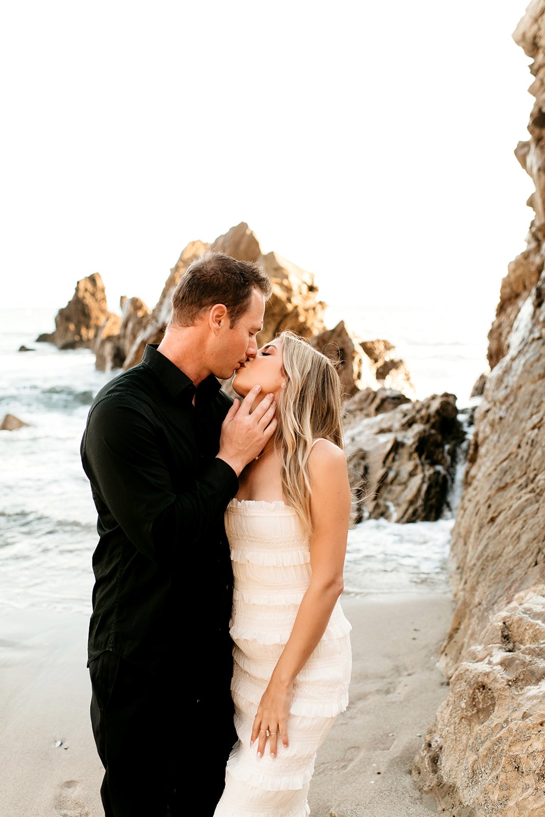 A couple sharing a kiss on the beach near rocky formations during sunset.
