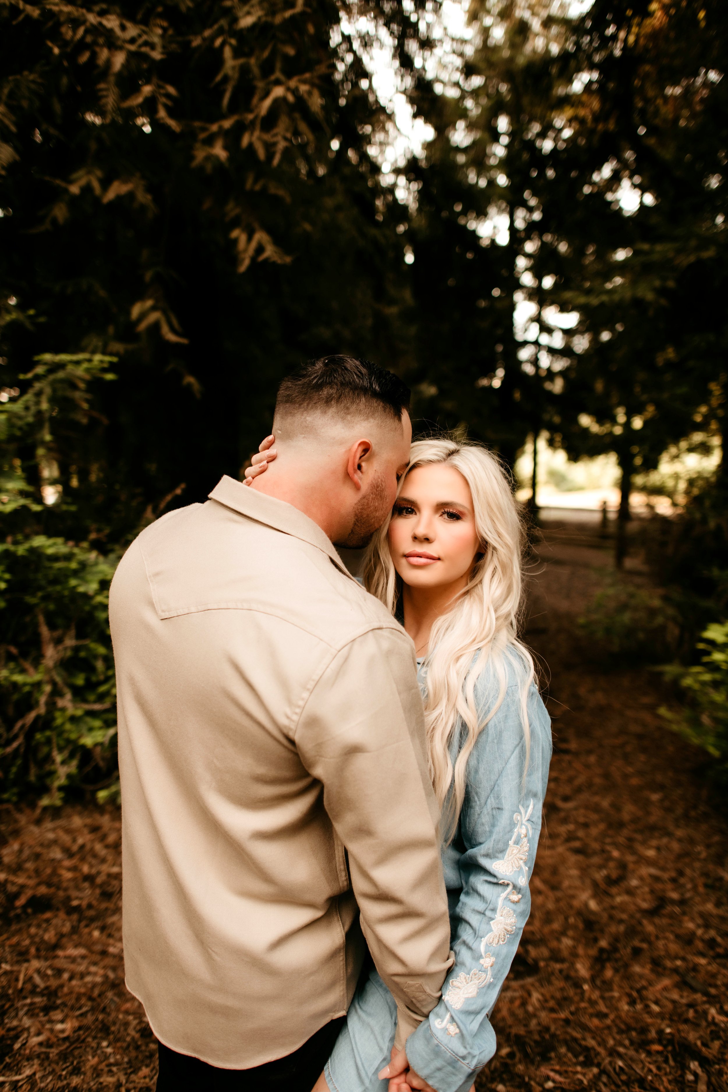 A man and woman standing outdoors in a wooded area, with the man leaning in to kiss the woman on the forehead. The woman has long, wavy blonde hair and is wearing a light blue denim jacket with floral embroidery, while the man has short dark hair and