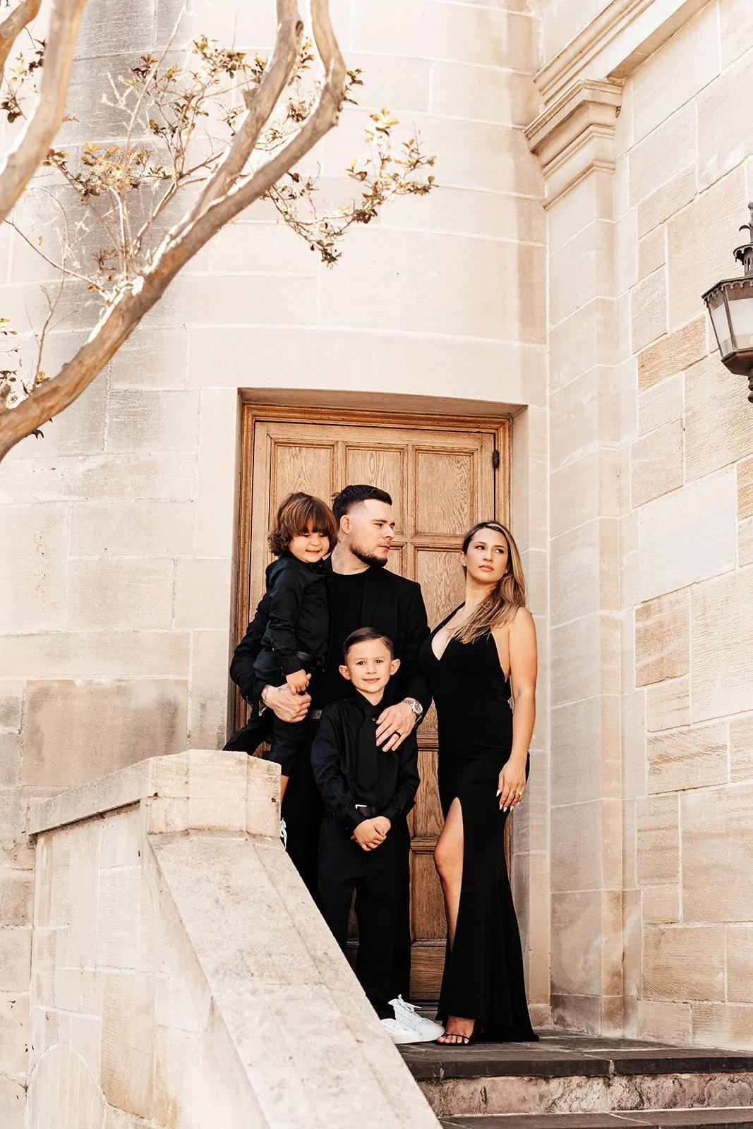 A family of four dressed in black standing on steps in front of a wooden door outside a beige stone building, with a tree and a lantern visible.