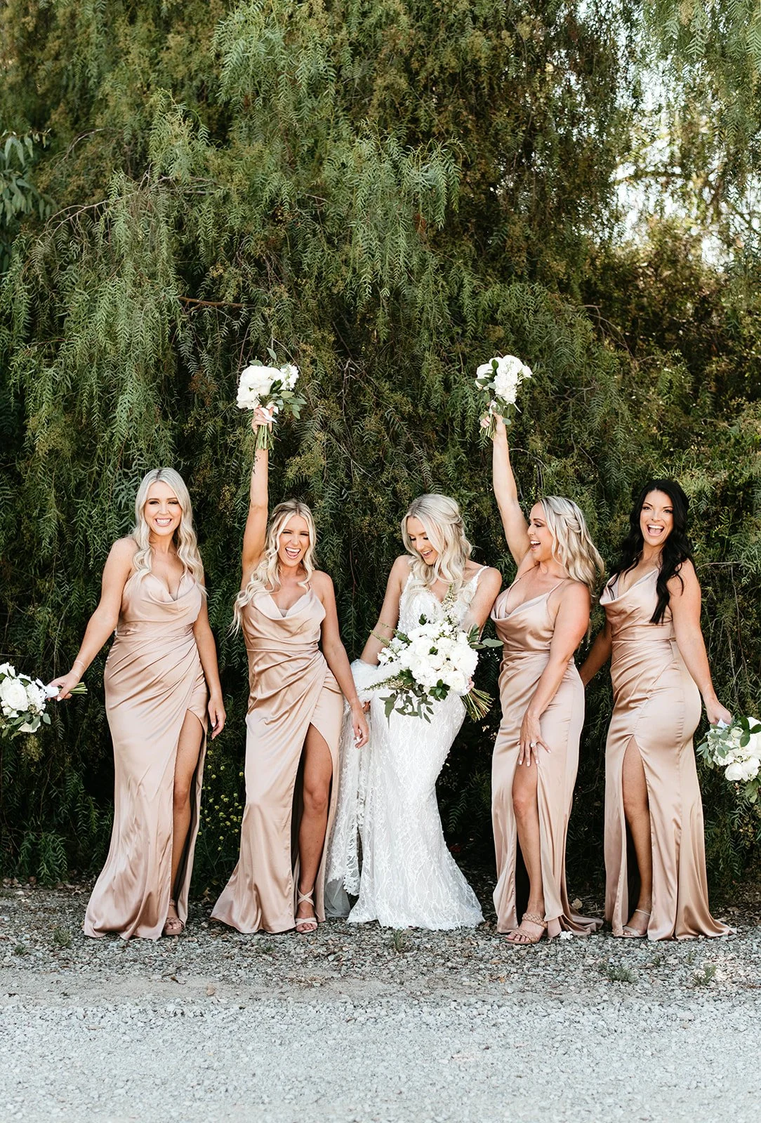 A group of five women, including a bride and four bridesmaids, happily celebrating outdoors against a lush green tree background. The bride is holding a large bouquet of white flowers, and the bridesmaids are holding smaller bouquets. Two bridesmaids
