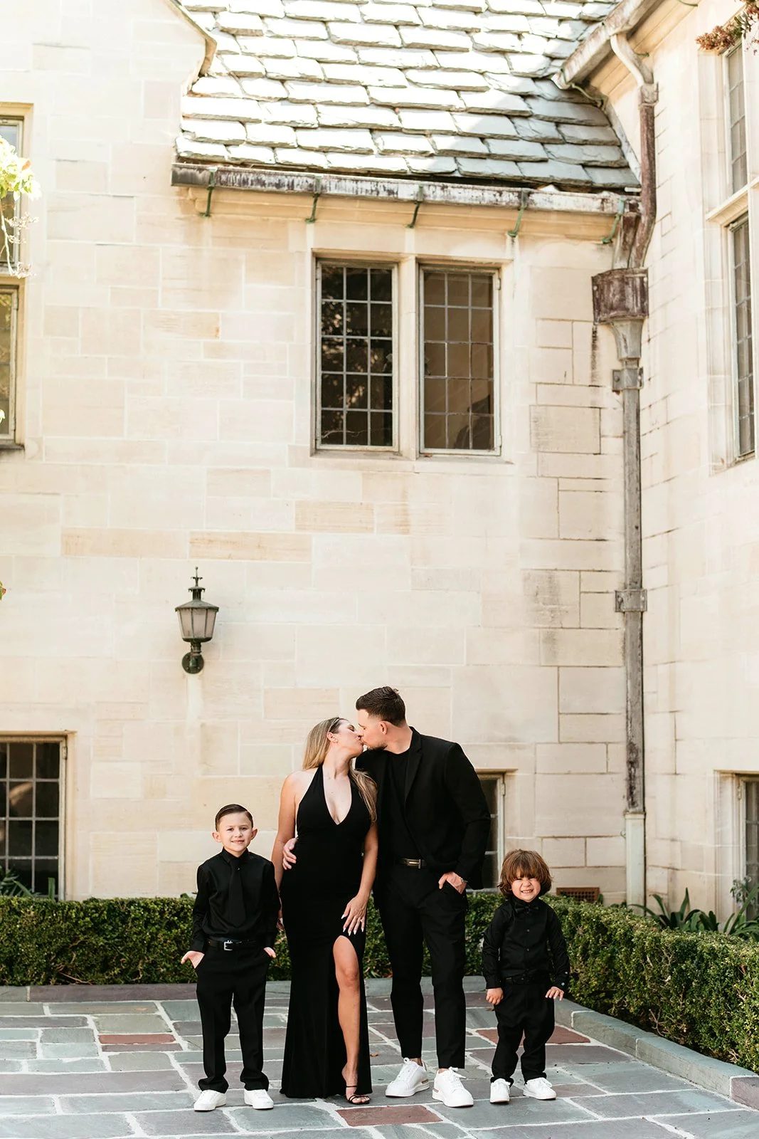 A family of four dressed in black, including a woman in a black gown and a man in a black suit, standing outdoors and sharing a kiss, with two young boys standing beside them, against the backdrop of an elegant stone building.