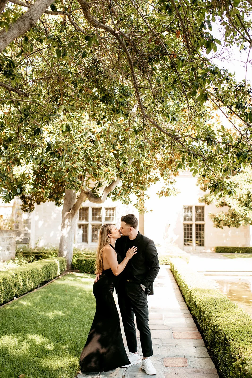 A couple dressed in black in a romantic pose, standing on a stone pathway under a large tree with green leaves, in front of a building with tall windows.