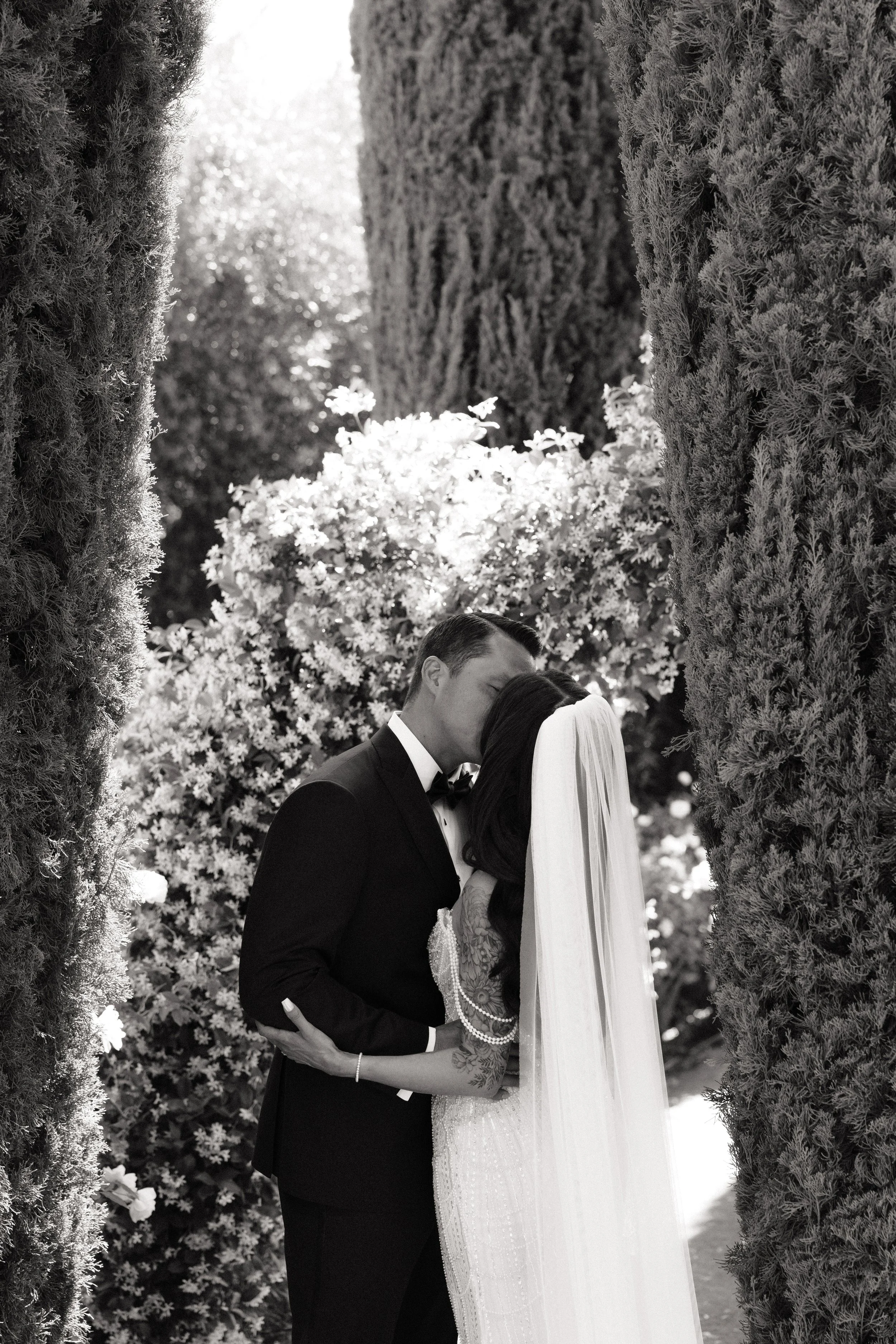 A black and white photo of a bride and groom kissing under an outdoor archway decorated with trees and flowers.
