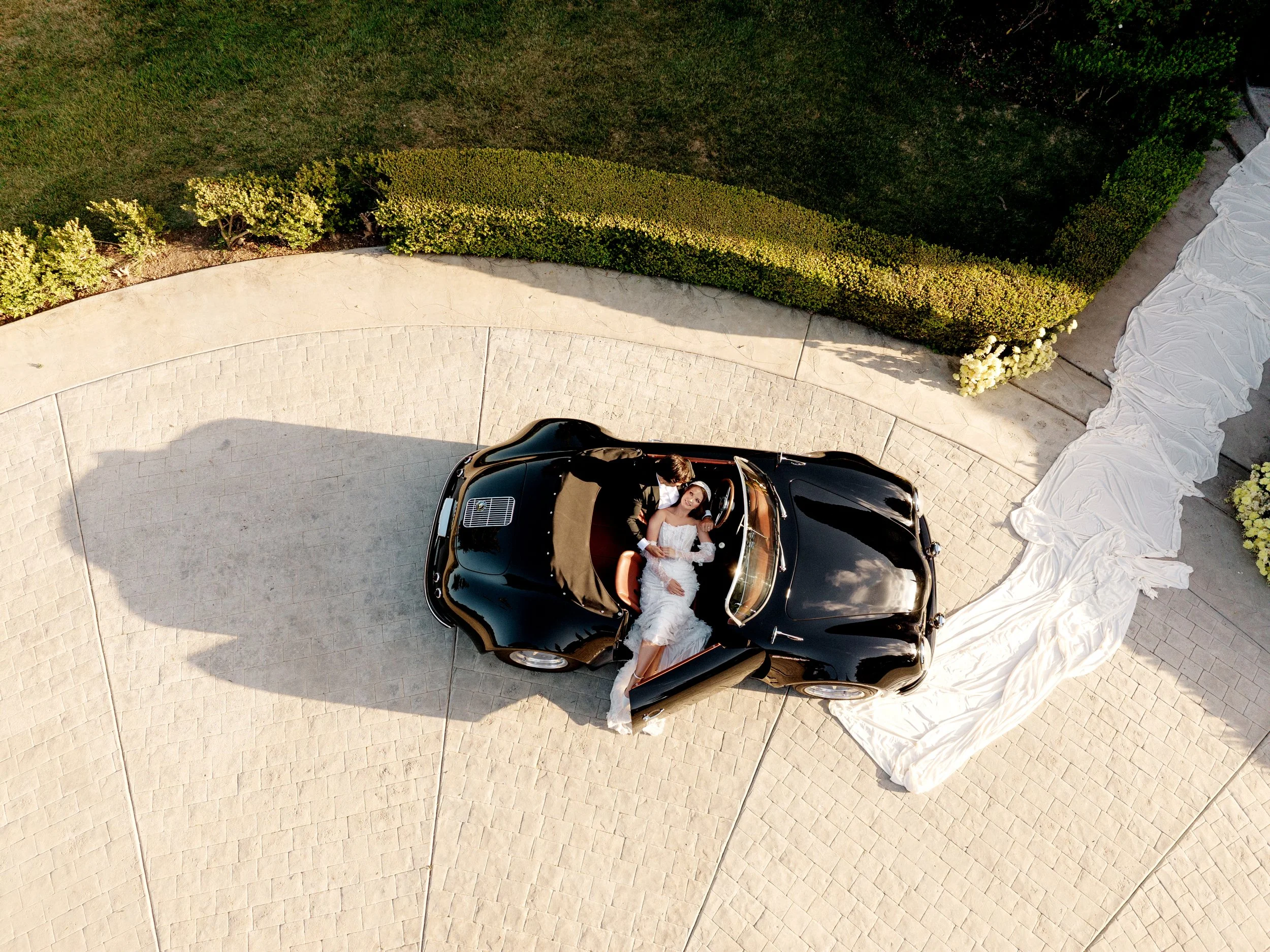 A bride and groom sitting in a black convertible car with a white cloth covering the backseat, taken from an aerial view. The bride is wearing a white wedding dress and the groom is dressed in a dark suit. They are smiling and looking upward.