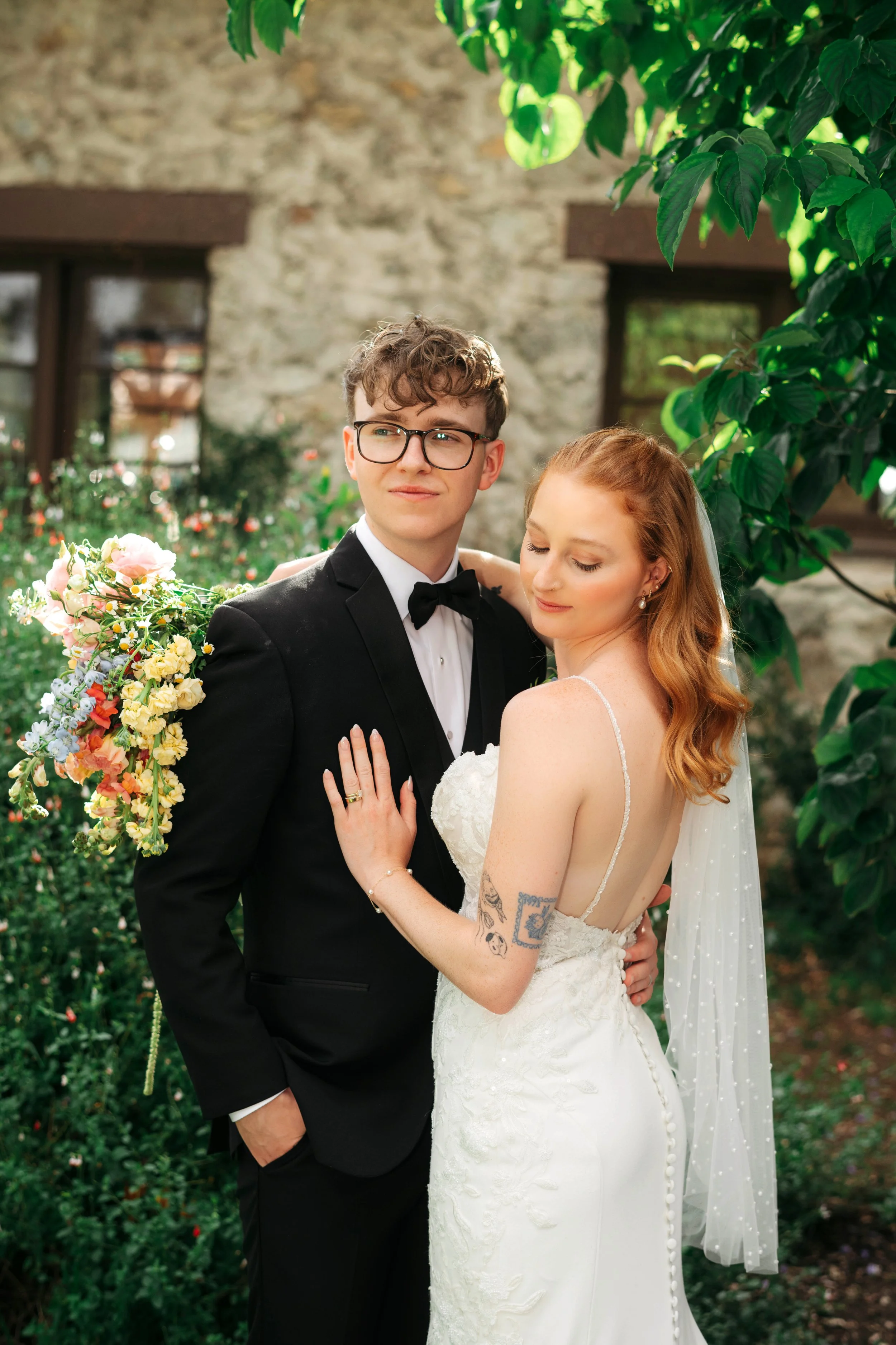 A happily married couple is posing outdoors for a wedding photo, with the groom wearing a tuxedo and the bride in a wedding dress, surrounded by greenery and a stone building in the background.