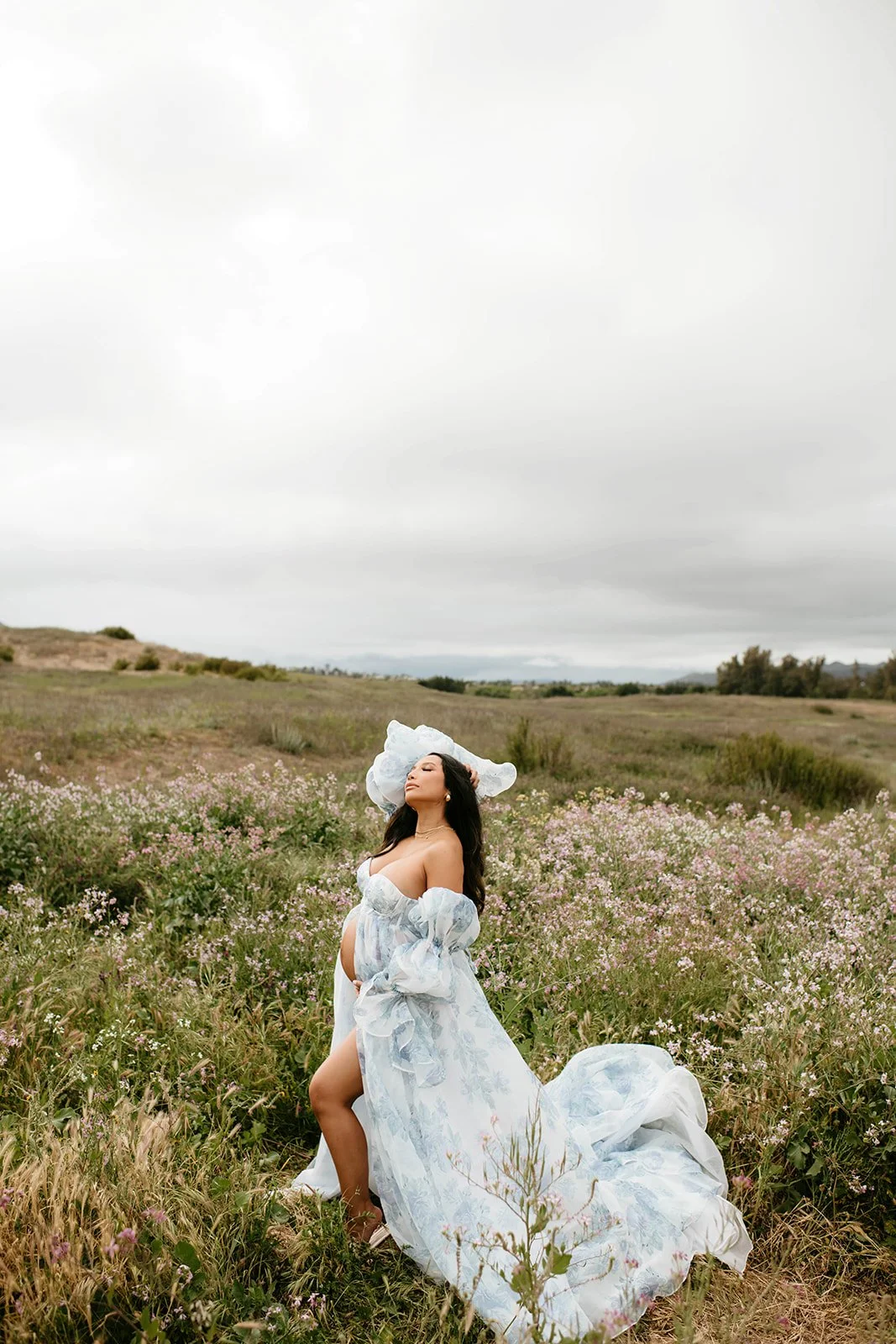 Pregnant woman in floral gown posing in a field of pink and purple flowers with overcast sky in the background.