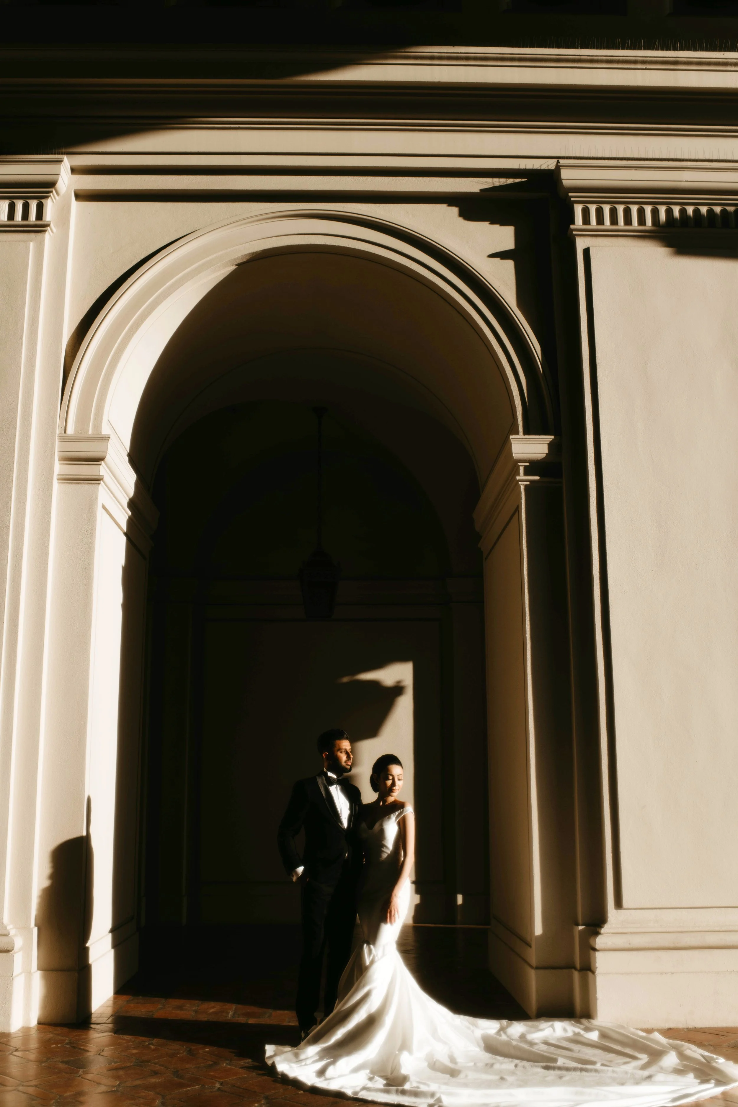 A bride and groom standing together under an archway in a sunlit architectural setting, with dramatic shadows. The bride is wearing a white gown with a long train, and the groom is in a black tuxedo. The sunlight highlights their faces and the elegan
