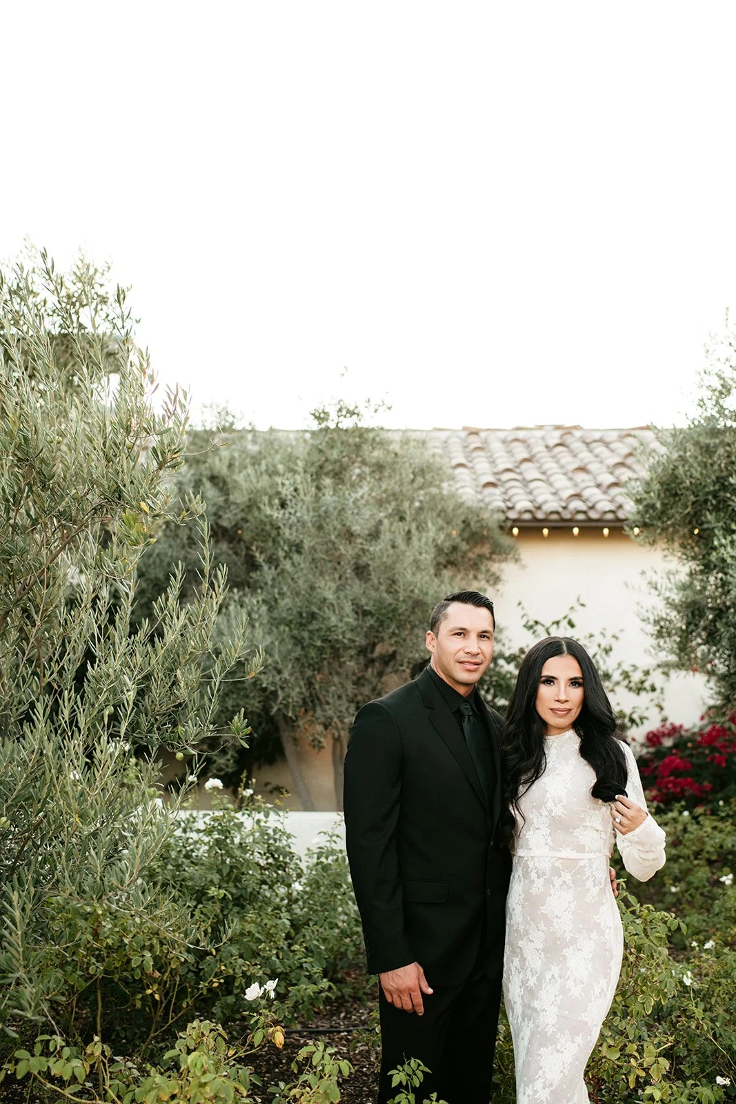 A couple dressed in formal attire standing in a lush garden with greenery and flowers, with a house with a tiled roof in the background.