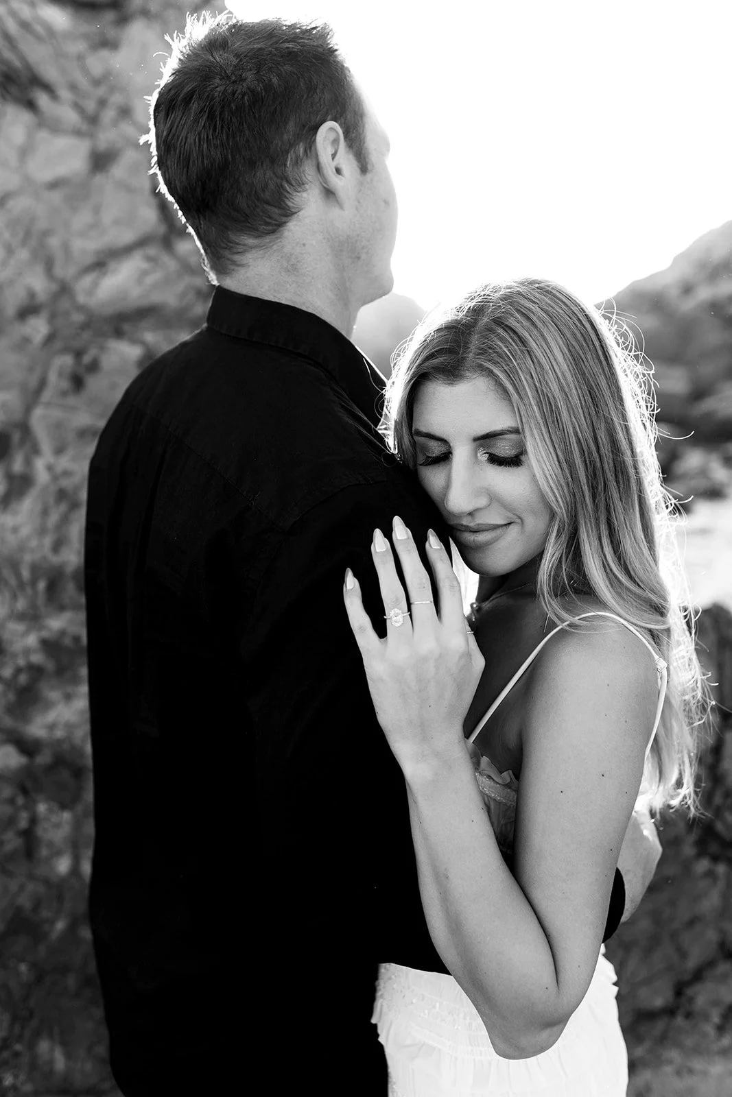 A black and white photo of a man and woman embracing outdoors. The woman has her eyes closed and is wearing a ring on her finger, with her hand resting on the man's shoulder. The man is facing away with his head turned slightly to the side. Sunlight 