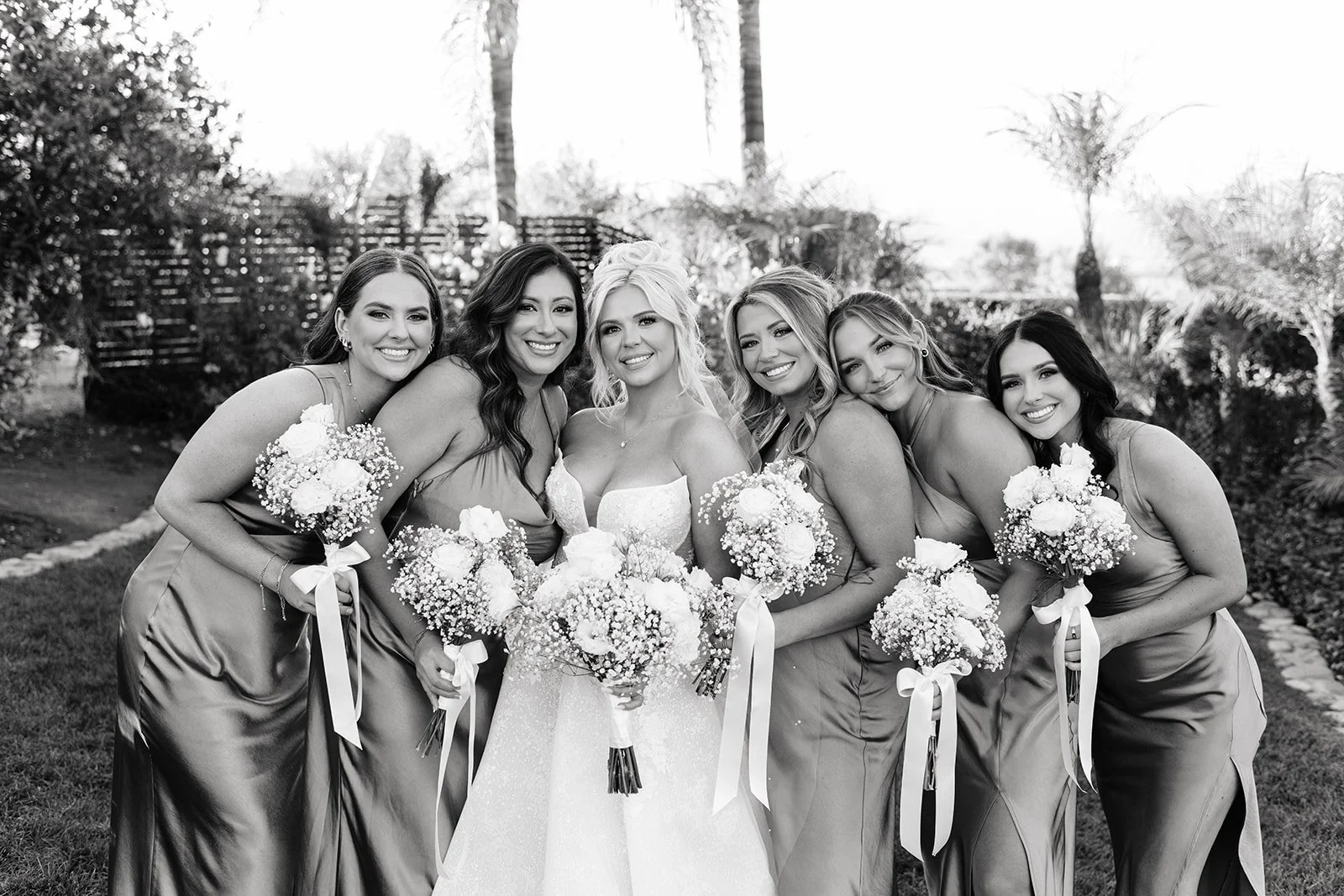 Black and white photo of a bride with five bridesmaids, all smiling and holding bouquets, outdoors with trees in the background.
