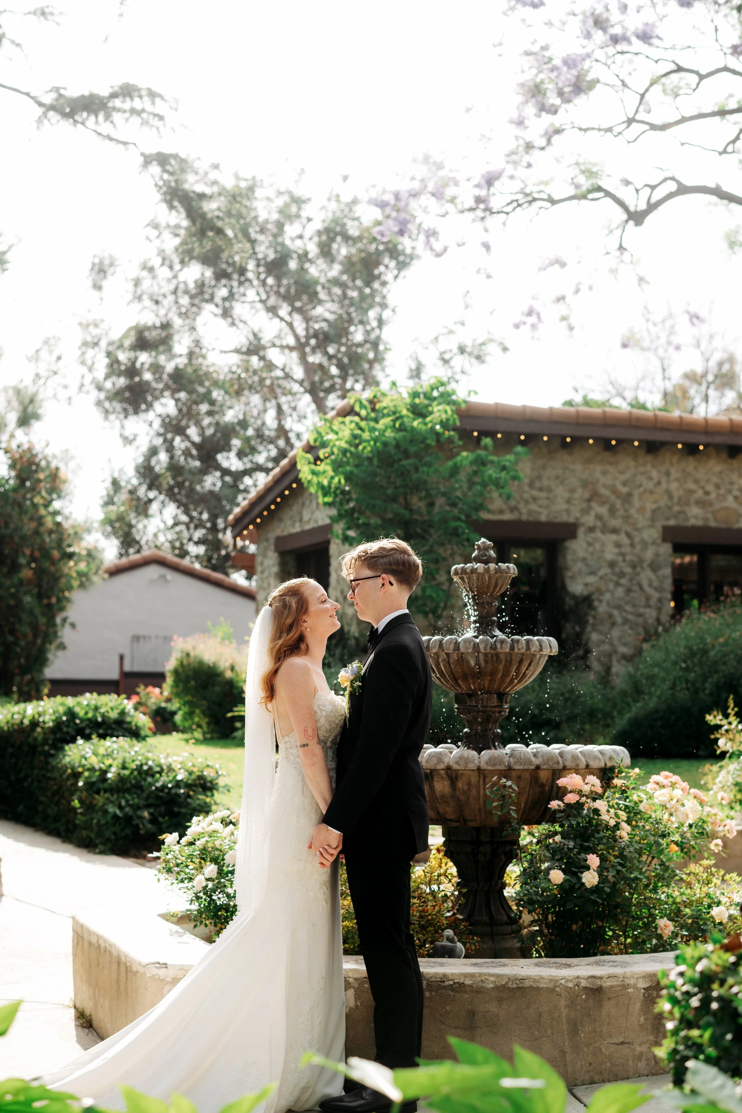 A bride and groom hold hands and look into each other's eyes in front of a fountain during their wedding ceremony outdoors, surrounded by greenery and colorful flowers.