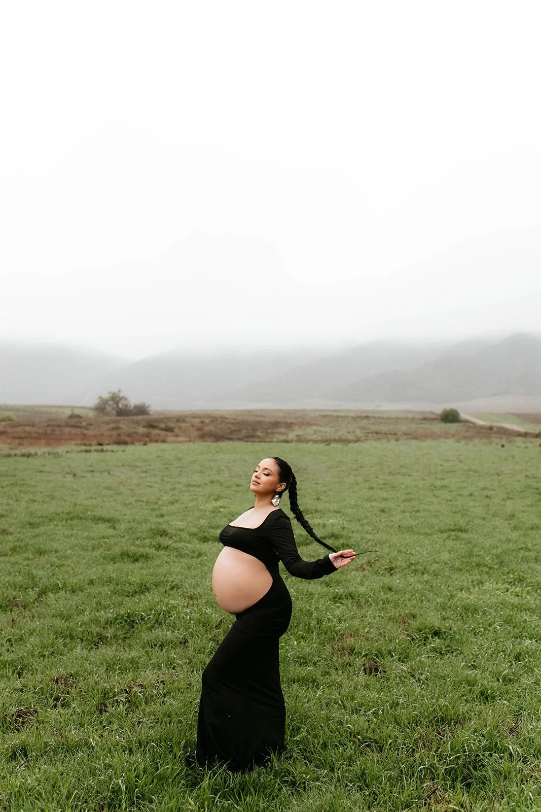 A pregnant woman in a black dress standing in a grassy field with mountains and cloudy sky in the background, holding her braid and smiling gently.
