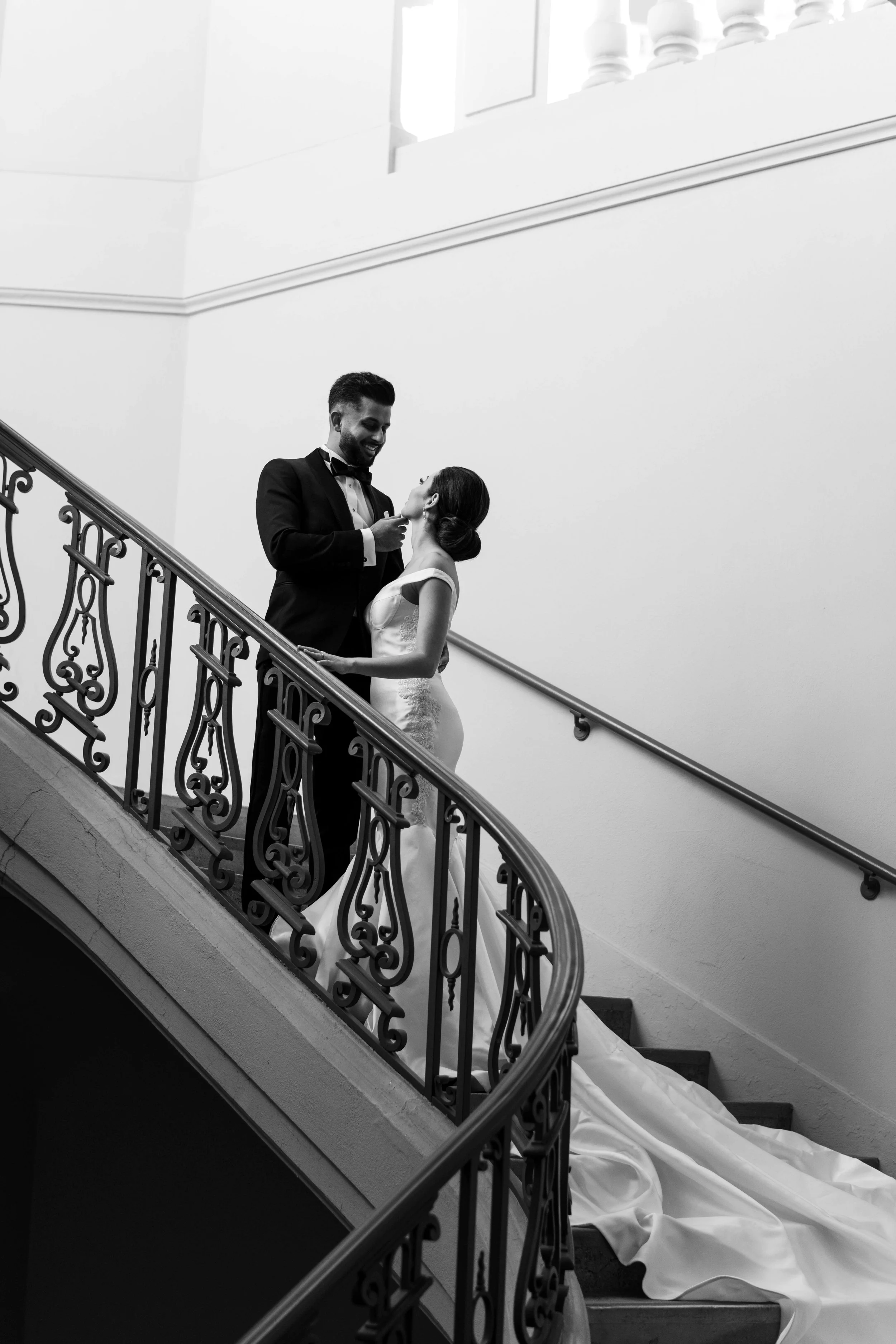 Black and white photo of a bride and groom standing on a staircase, looking at each other, with the bride's wedding dress trailing down the stairs.