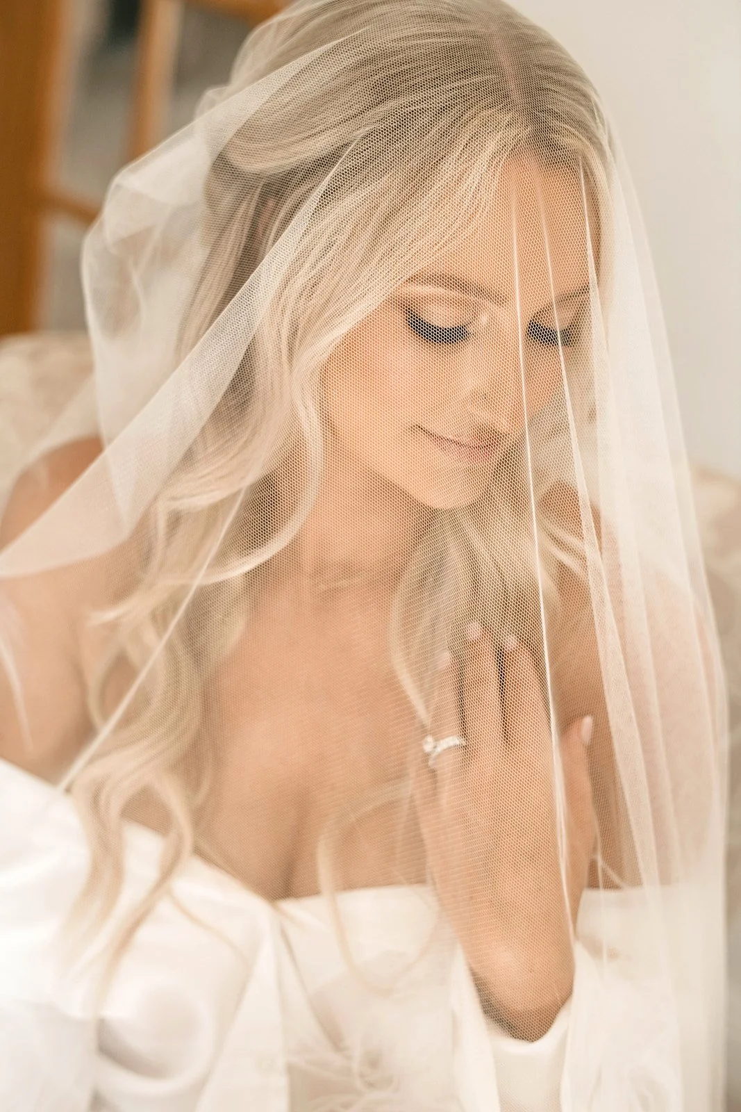 A bride with long blonde hair and a veil over her face, smiling softly with eyes closed.