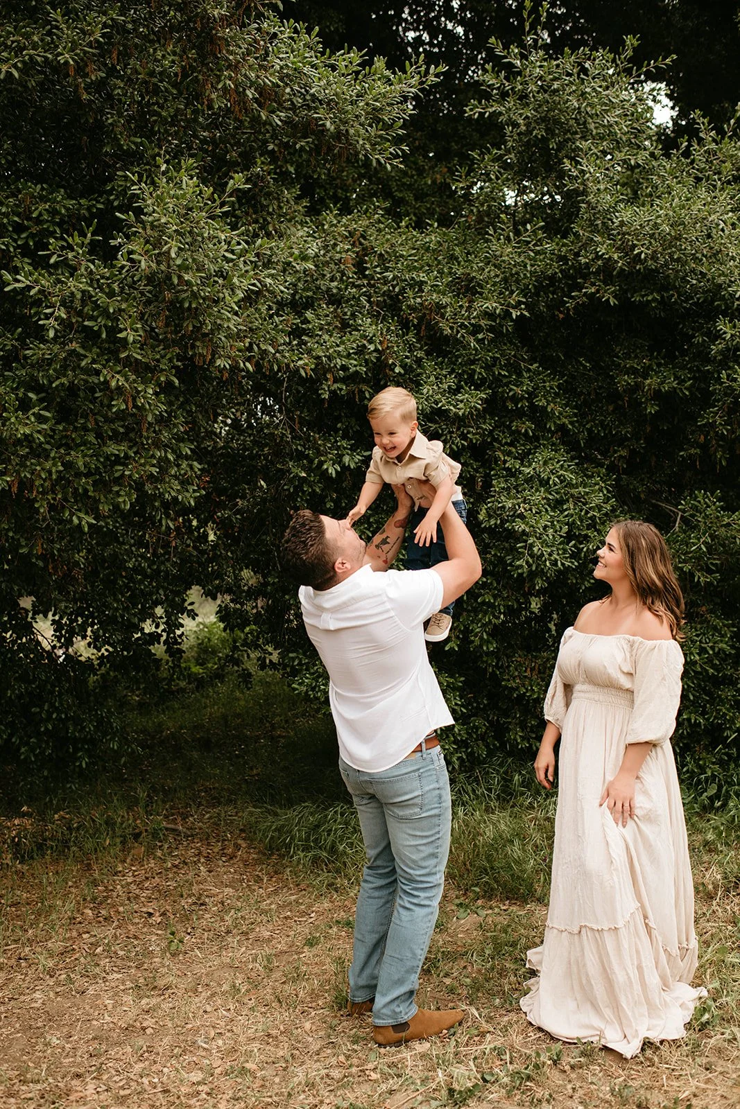 A family outdoors enjoying time together. A man is lifting a young boy into the air while a woman, standing nearby, watches and smiles. They are surrounded by green trees and grass.