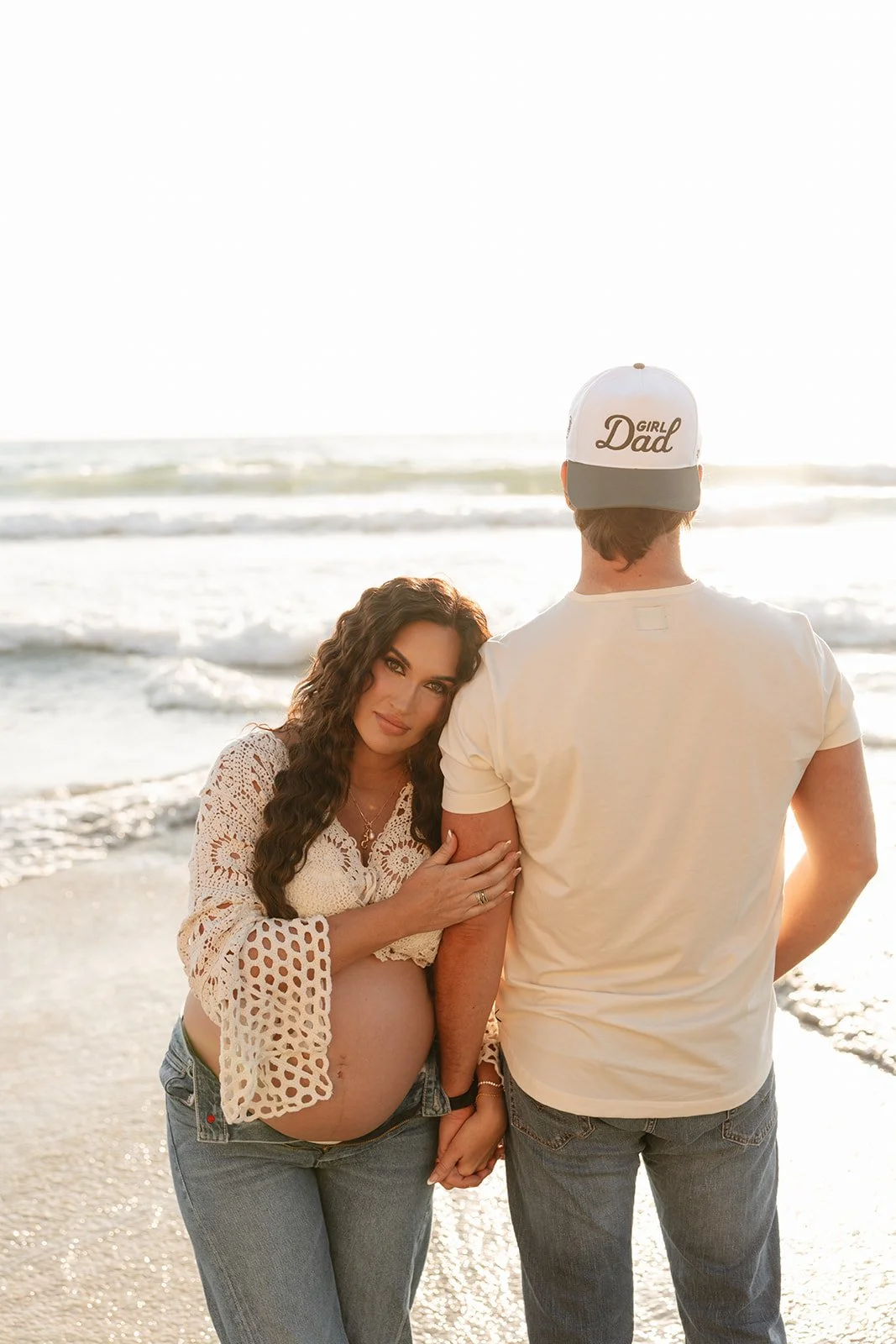 A pregnant woman with long curly hair in a crochet top standing on the beach, holding hands with a man with a cap that says "Girl Dad," both facing the ocean at sunset.