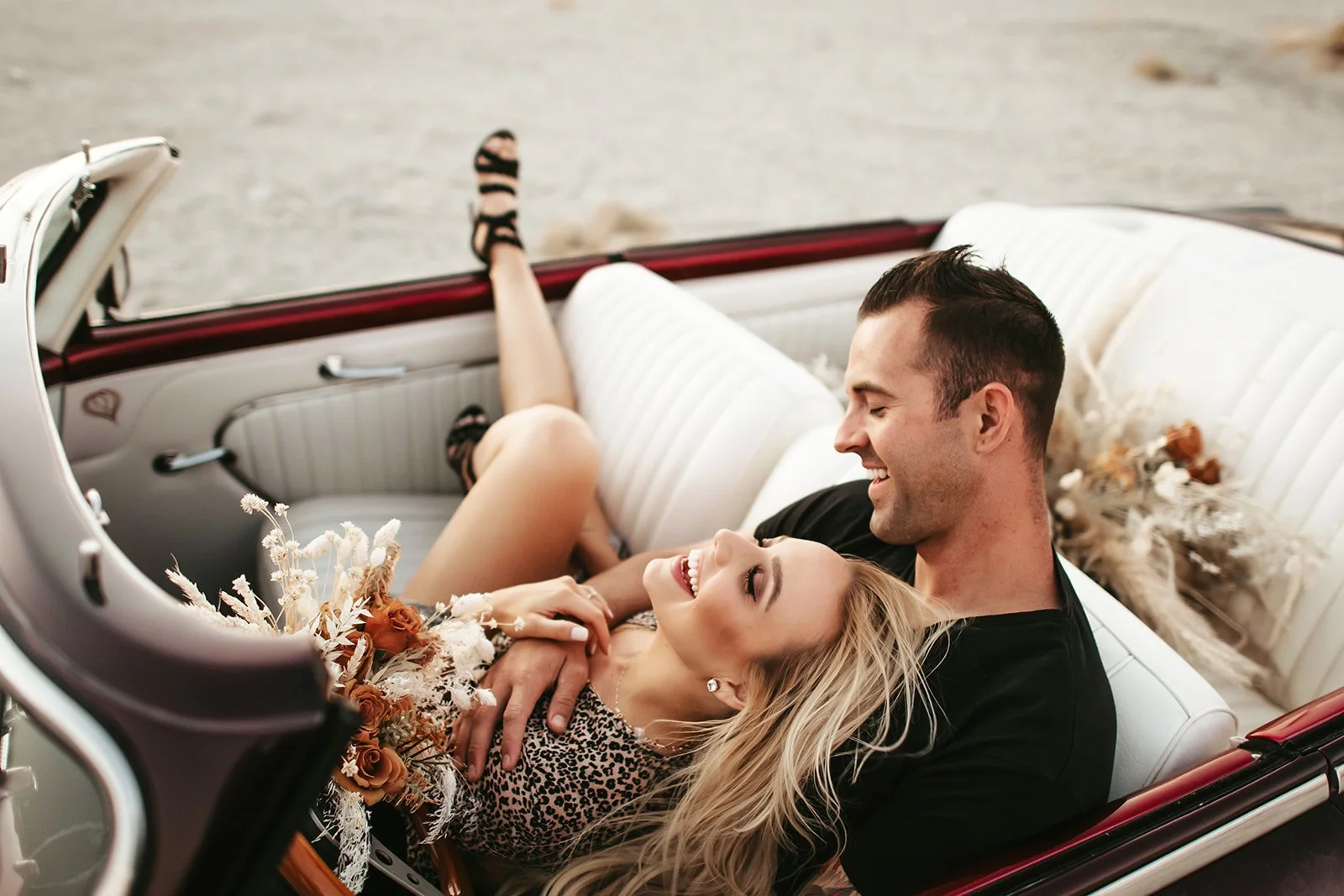 A happy couple sitting in a vintage car, smiling and enjoying each other's company, with the woman holding a bouquet of flowers.