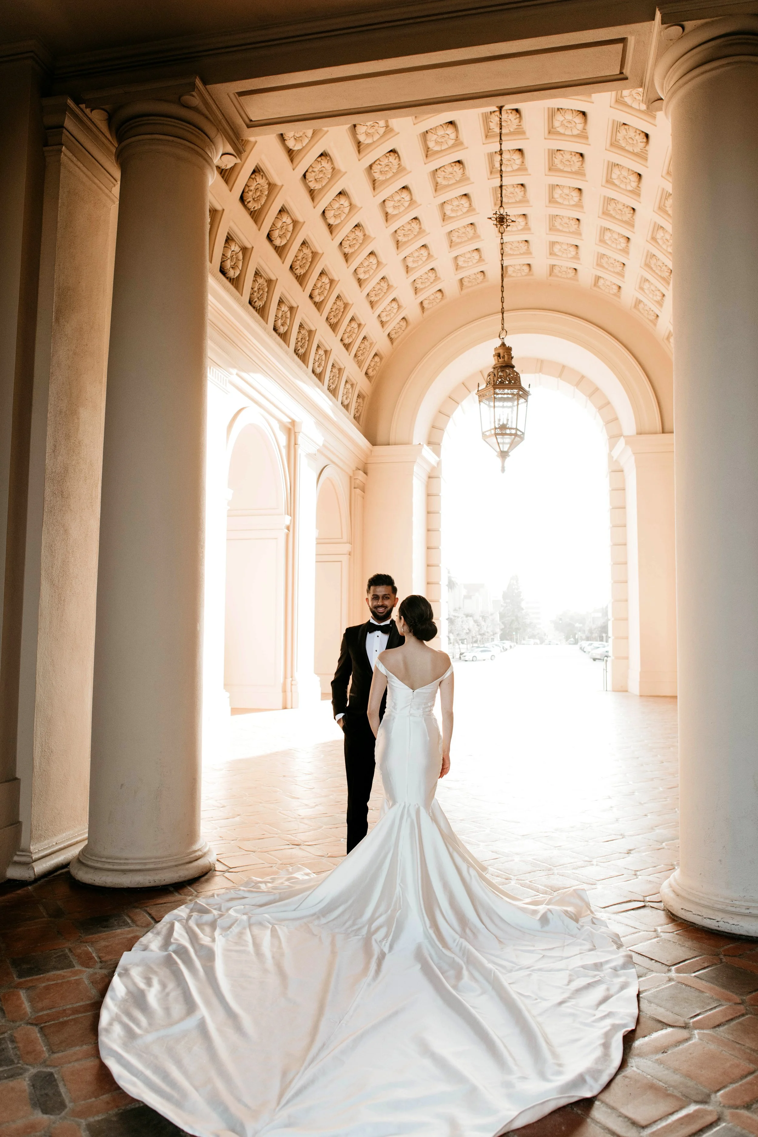A bride and groom standing under a decorative archway with columns, backlit by bright natural light, with the bride in a white gown with a long train and the groom in a tuxedo.