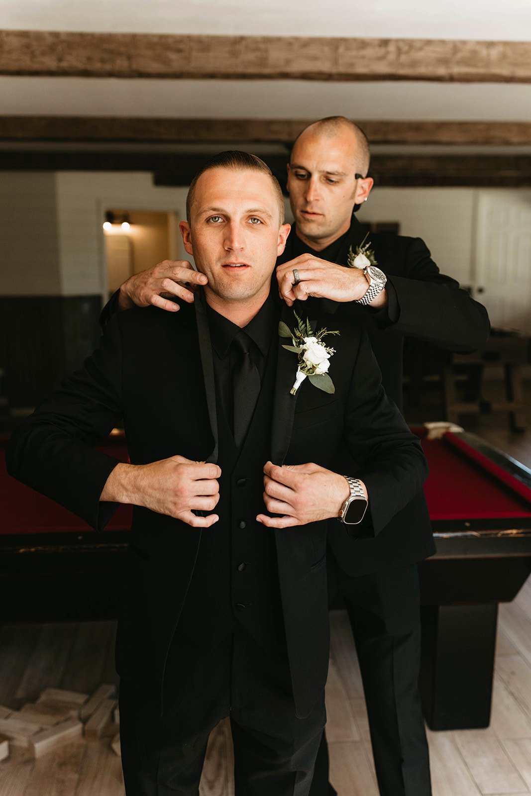 Two men dressed in black suits, with boutonnières, getting ready for a formal event. One man is adjusting his jacket, while the other assists by fixing his collar.