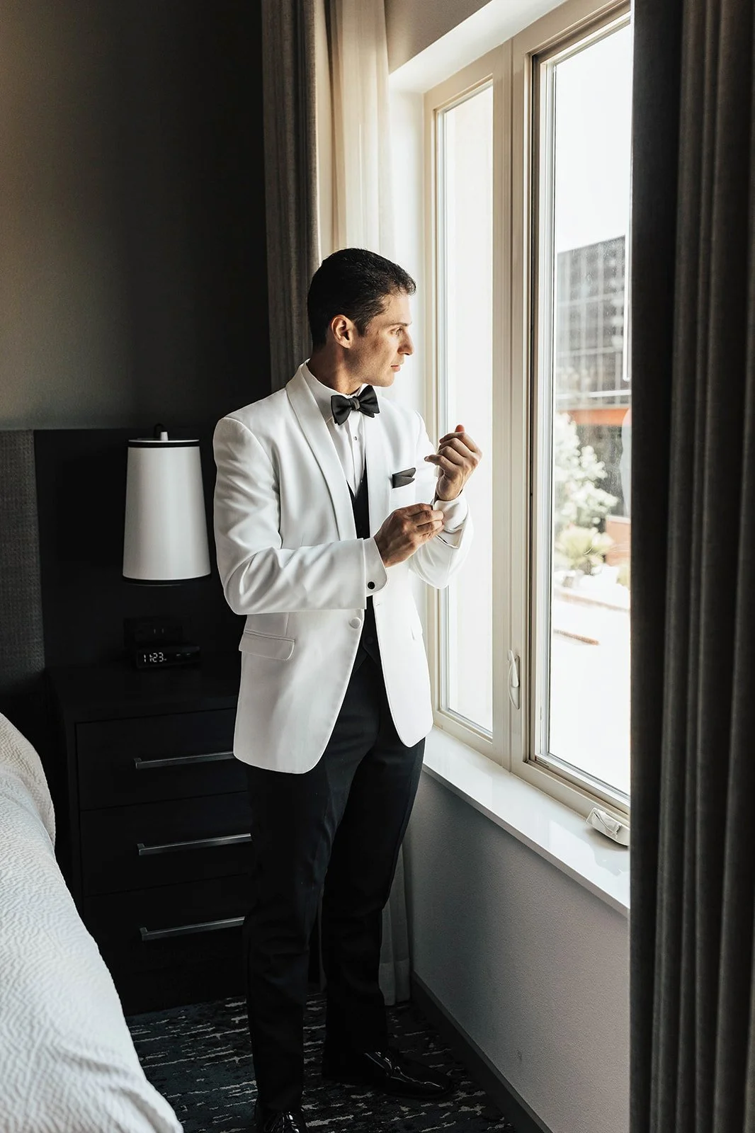 A man in a white tuxedo jacket, black pants, and a bow tie stands by a window, adjusting a cufflink, with a bedside table and lamp nearby.