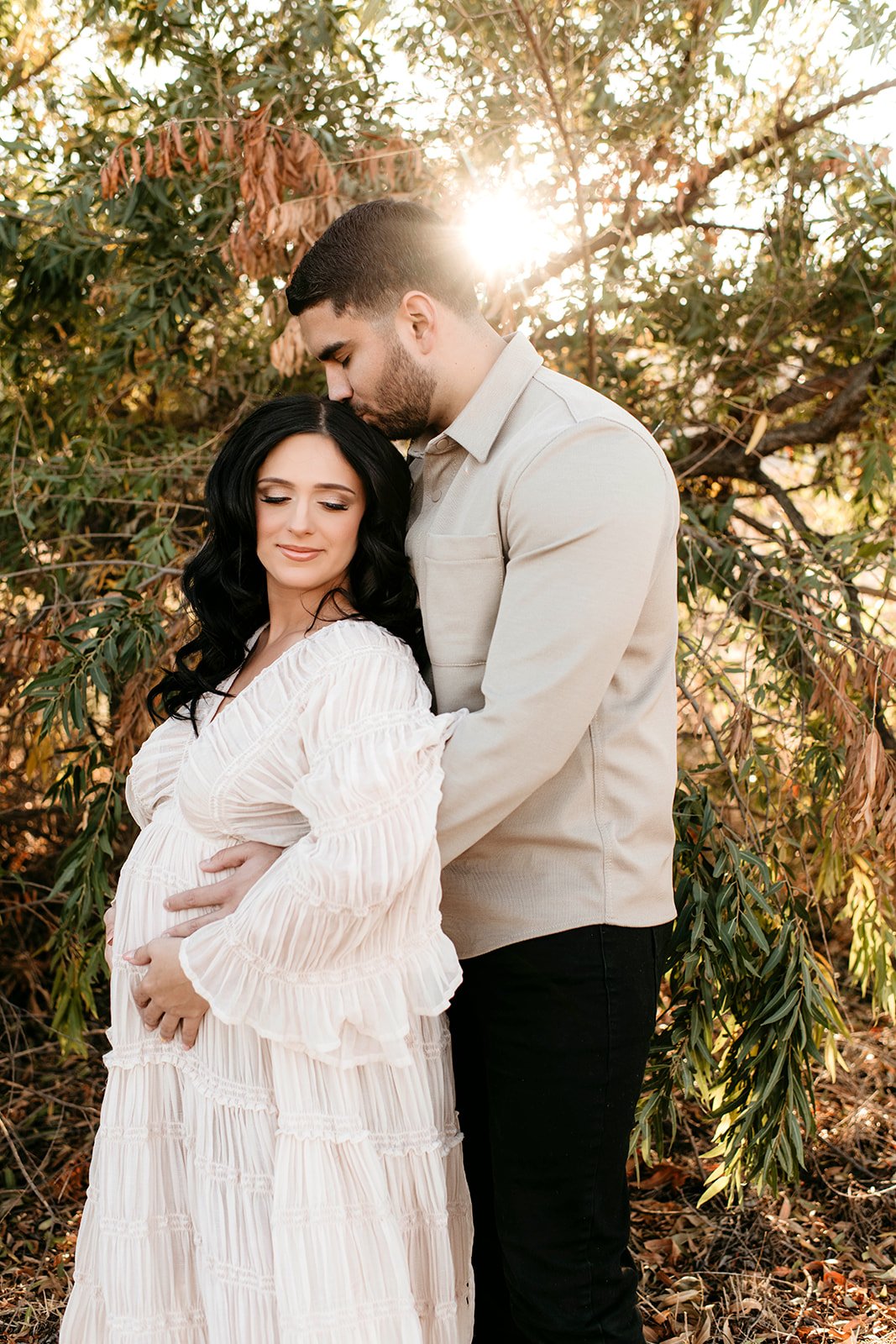 A pregnant woman with long black hair in a white dress, standing next to a man in a light-colored shirt in a natural outdoor setting with trees. The man is kissing her forehead, and she is smiling with her eyes closed. Sunlight filters through the tr