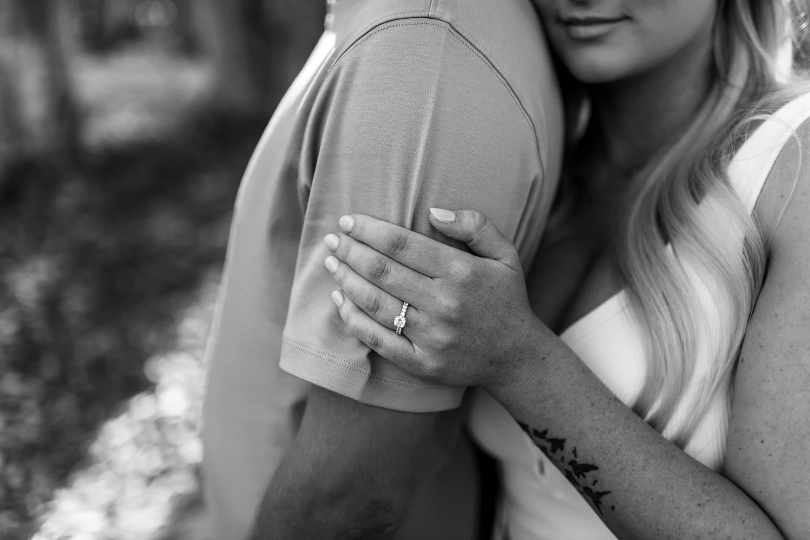 A woman is resting her hand on a man's shoulder, showing a ring on her finger, with her long blond hair and an engagement ring visible in black and white.
