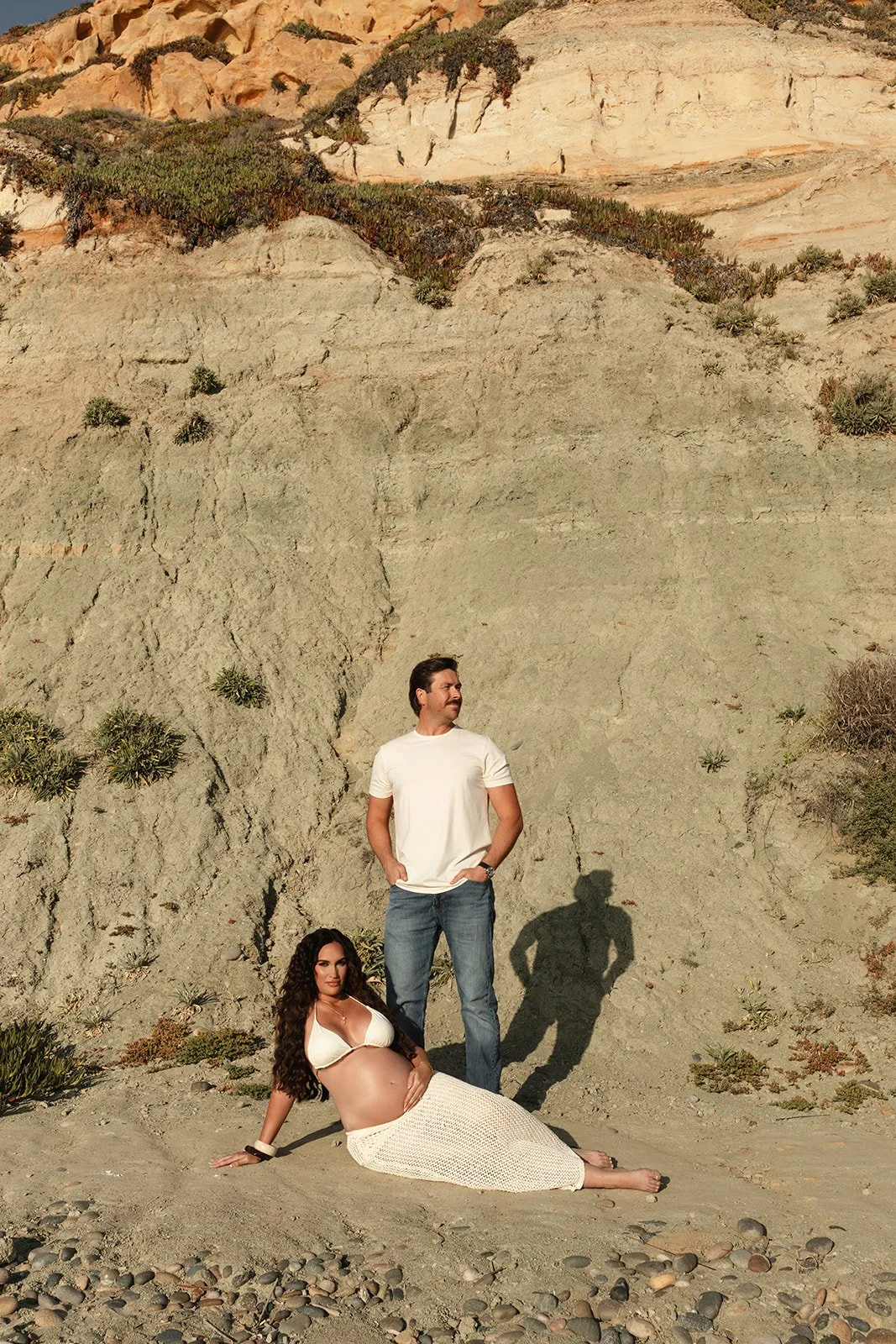 A man and a pregnant woman posing on a sandy beach in front of a rocky cliff with sparse vegetation, with the woman sitting on the ground and the man standing behind her.