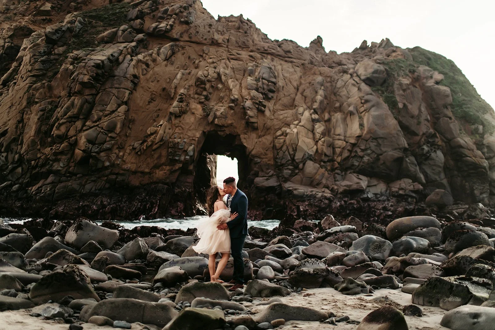 A couple dressed in formal attire, with the woman in a wedding dress and the man in a suit, standing on rocks on a beach, sharing a kiss in front of a natural rock arch formation.