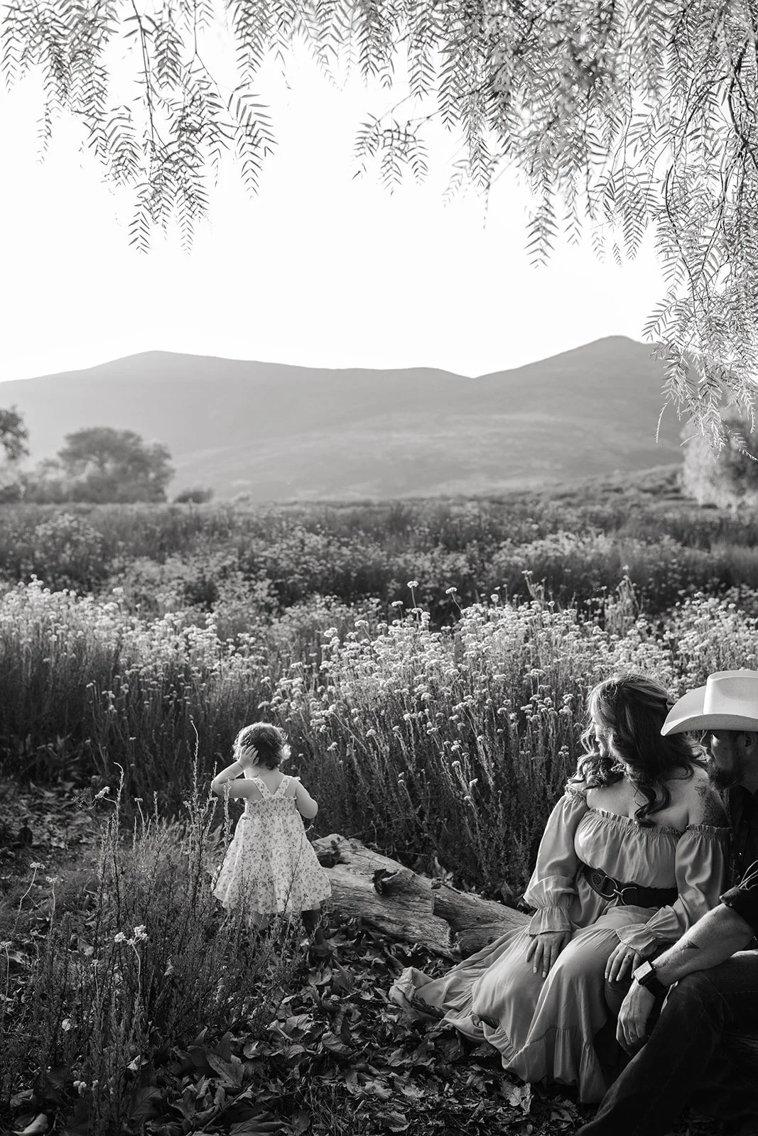 A black and white photo of a woman and a man sitting on a log in a field of flowers, with a young girl standing nearby looking at the landscape. The woman is wearing a dress with off-the-shoulder sleeves, and the man is wearing a cowboy hat. The back