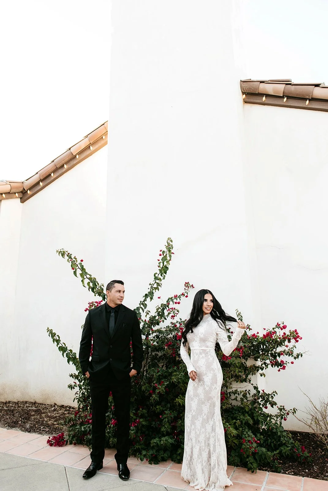 A man in a black suit and woman in a white lace dress standing outdoors in front of a wall with flowering bushes.