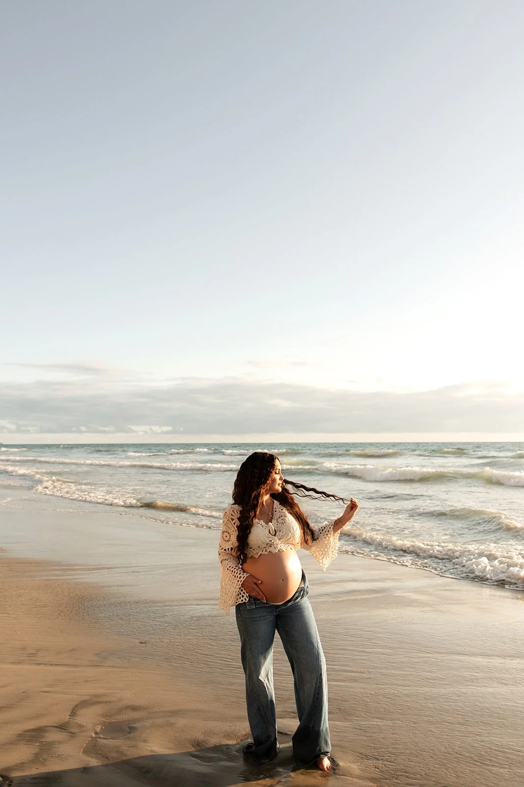 Pregnant woman standing on the beach with ocean waves in the background, wearing jeans and a beige crocheted top, holding her long hair and smiling.