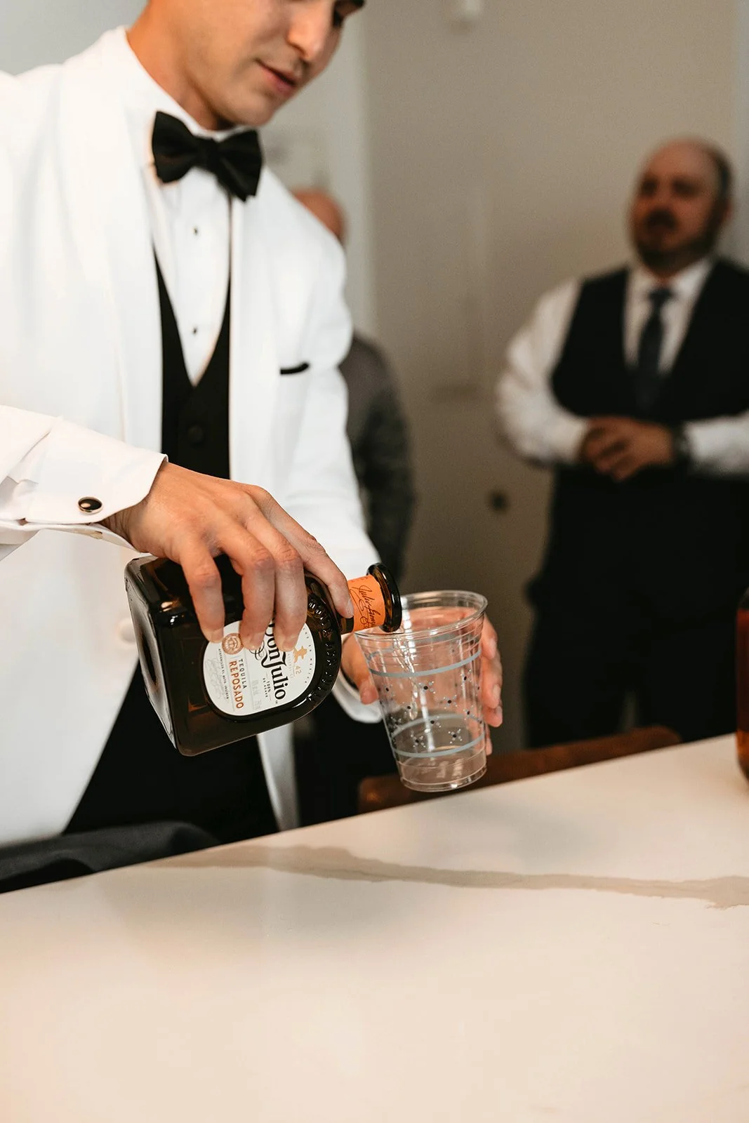 A man in a tuxedo and dress shirt pours a drink from a dark bottle into a clear plastic cup, with two men in vests and dress shirts watching in the background.